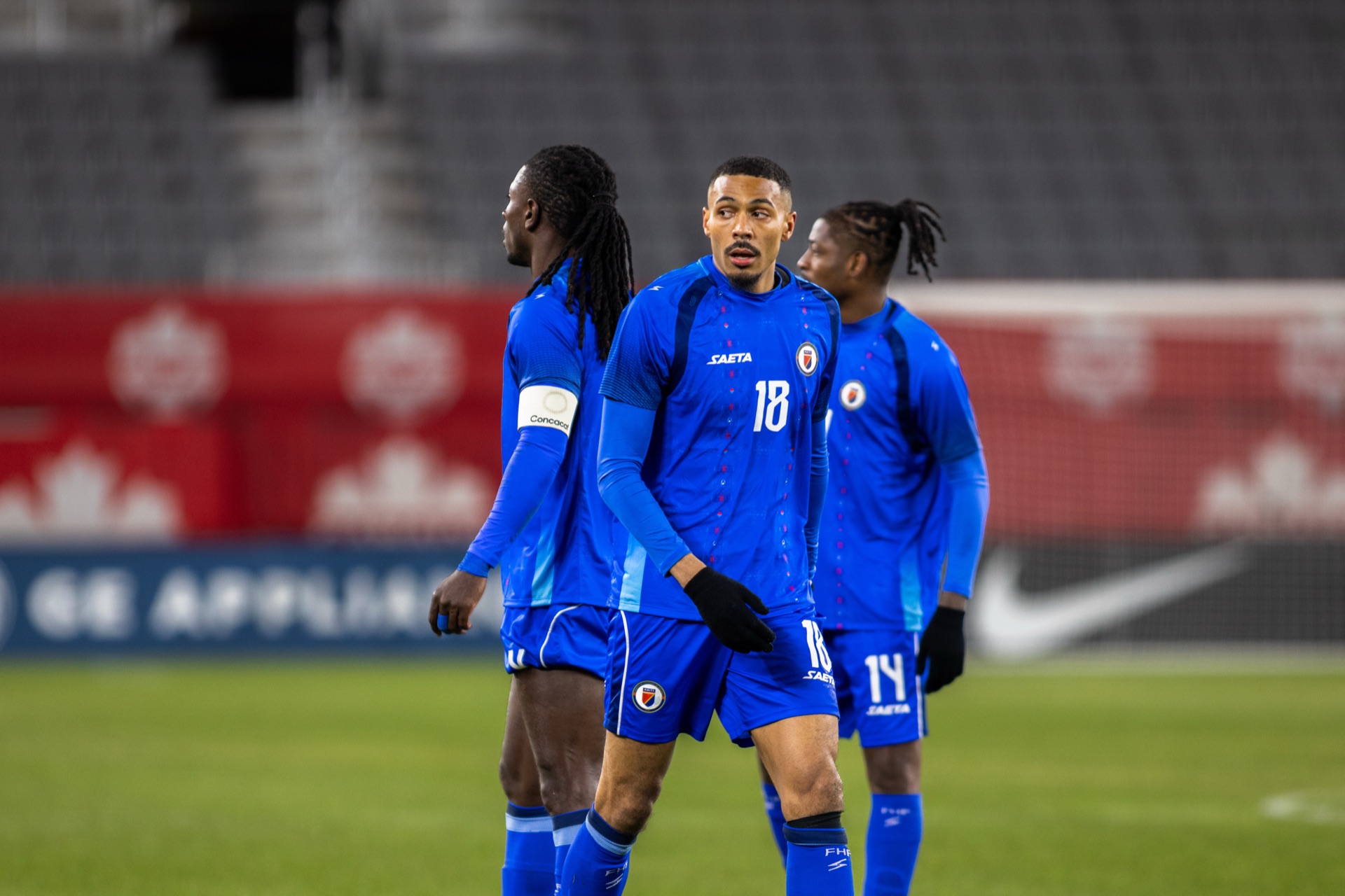 Isidor walking up the pitch between two Haiti teammates, red Canada Soccer boards behind them