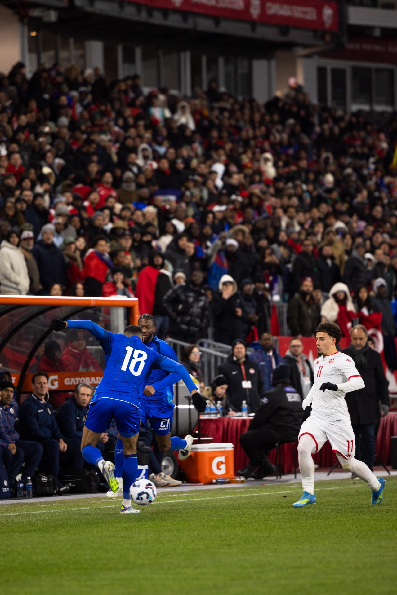 Isidor striking the ball from the touchline in front of the benches, a Tunisia player closing him down and a full stand behind