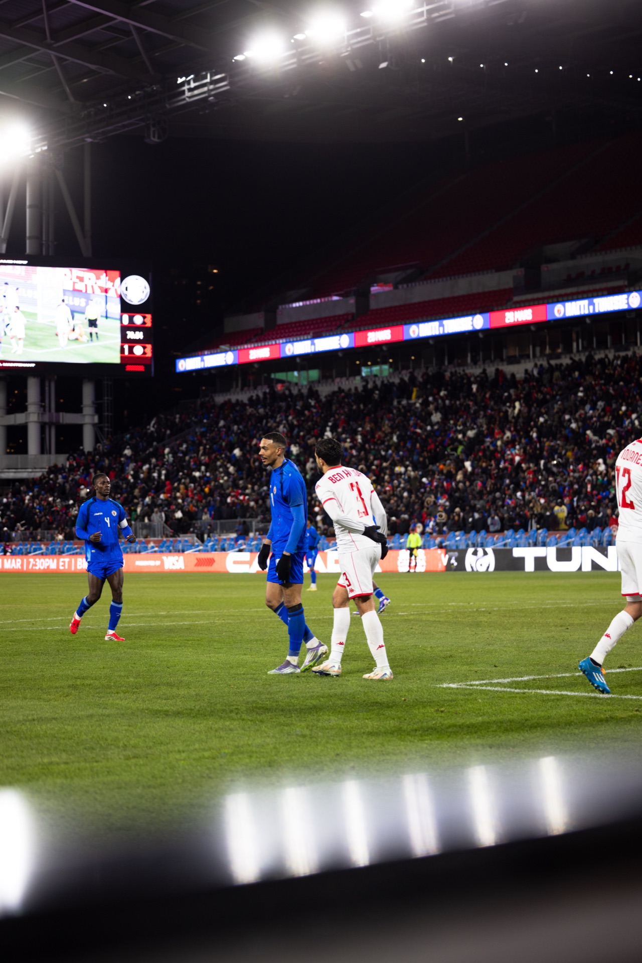 Wide view from pitch level — floodlights, the big screen and a full stand, with Isidor and two Tunisia players in the middle distance