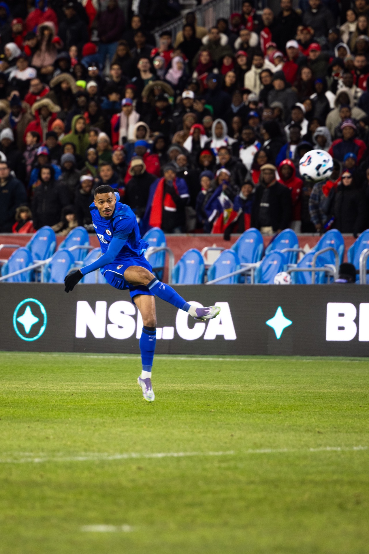 Isidor off the ground with his leg swung through a volley, the ball at head height and supporters behind the hoarding