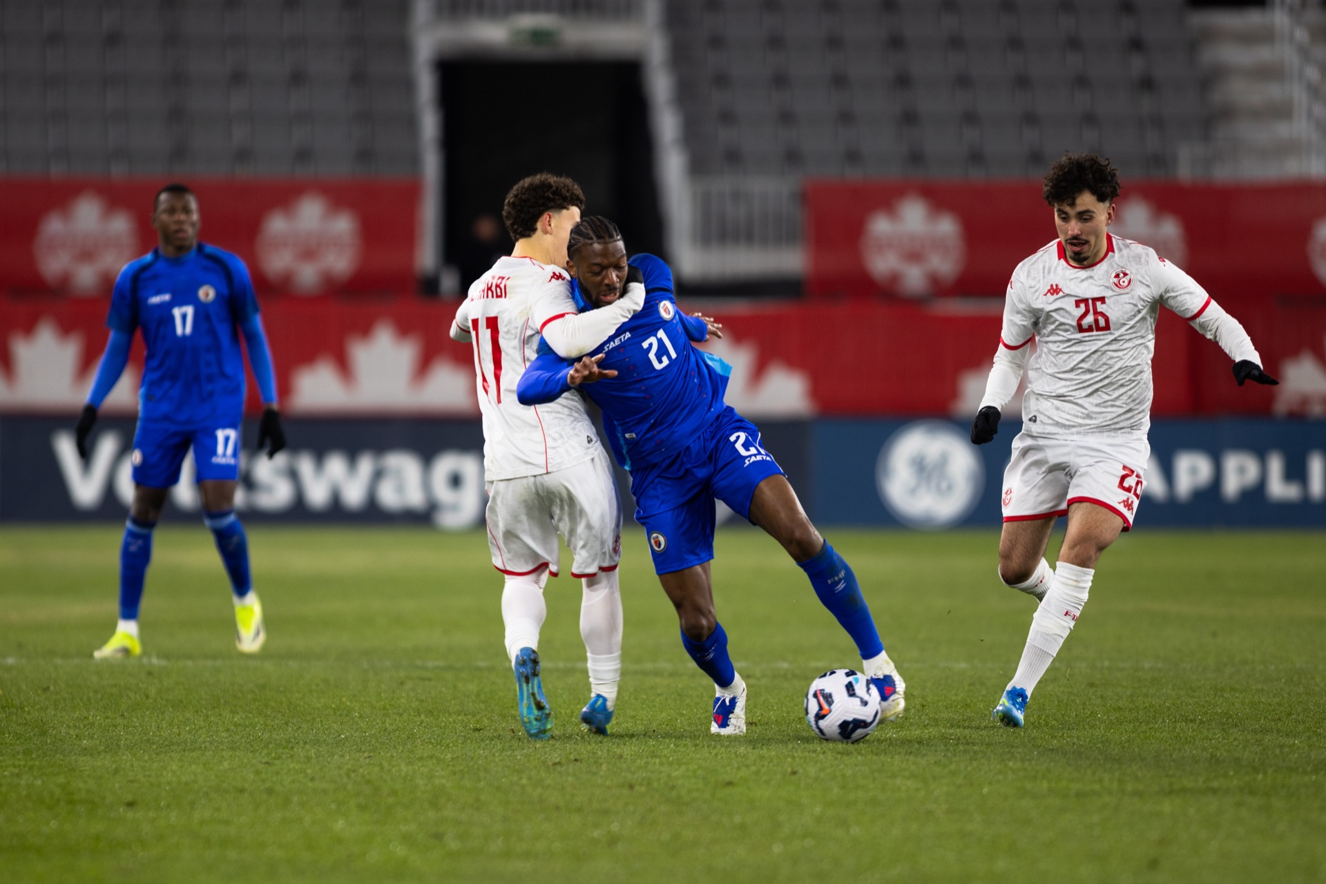 A Haiti player carrying the ball away from a Tunisia defender, a second Tunisia shirt blurred across the foreground