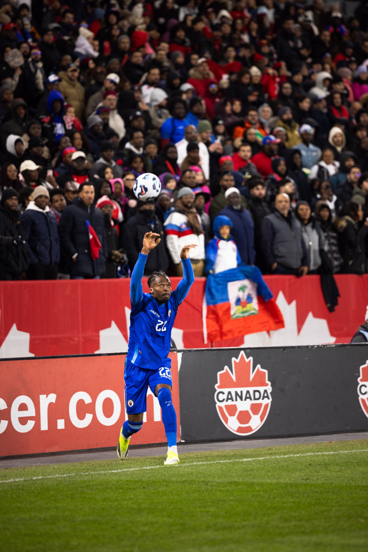 A Haiti player taking a throw-in with the ball above his head, a Haitian flag draped over the barrier in the crowd behind