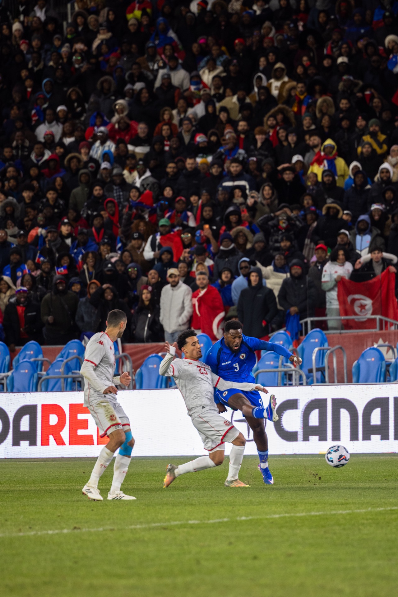 Haiti's number 9 held off by two Tunisia players, a Tunisian flag raised in the stand behind them