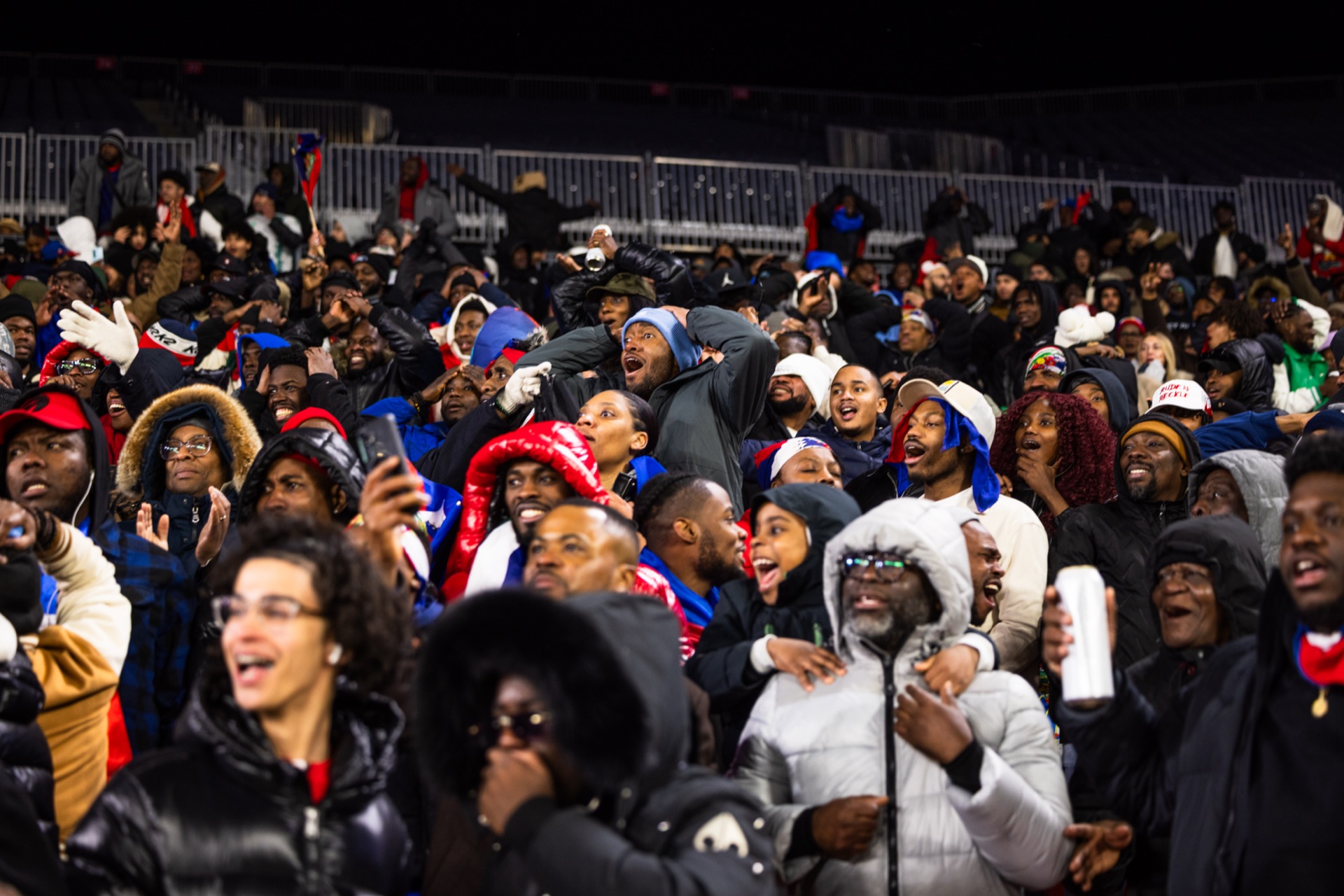 Haiti supporters in the stand on their feet in winter coats, shouting, phones raised