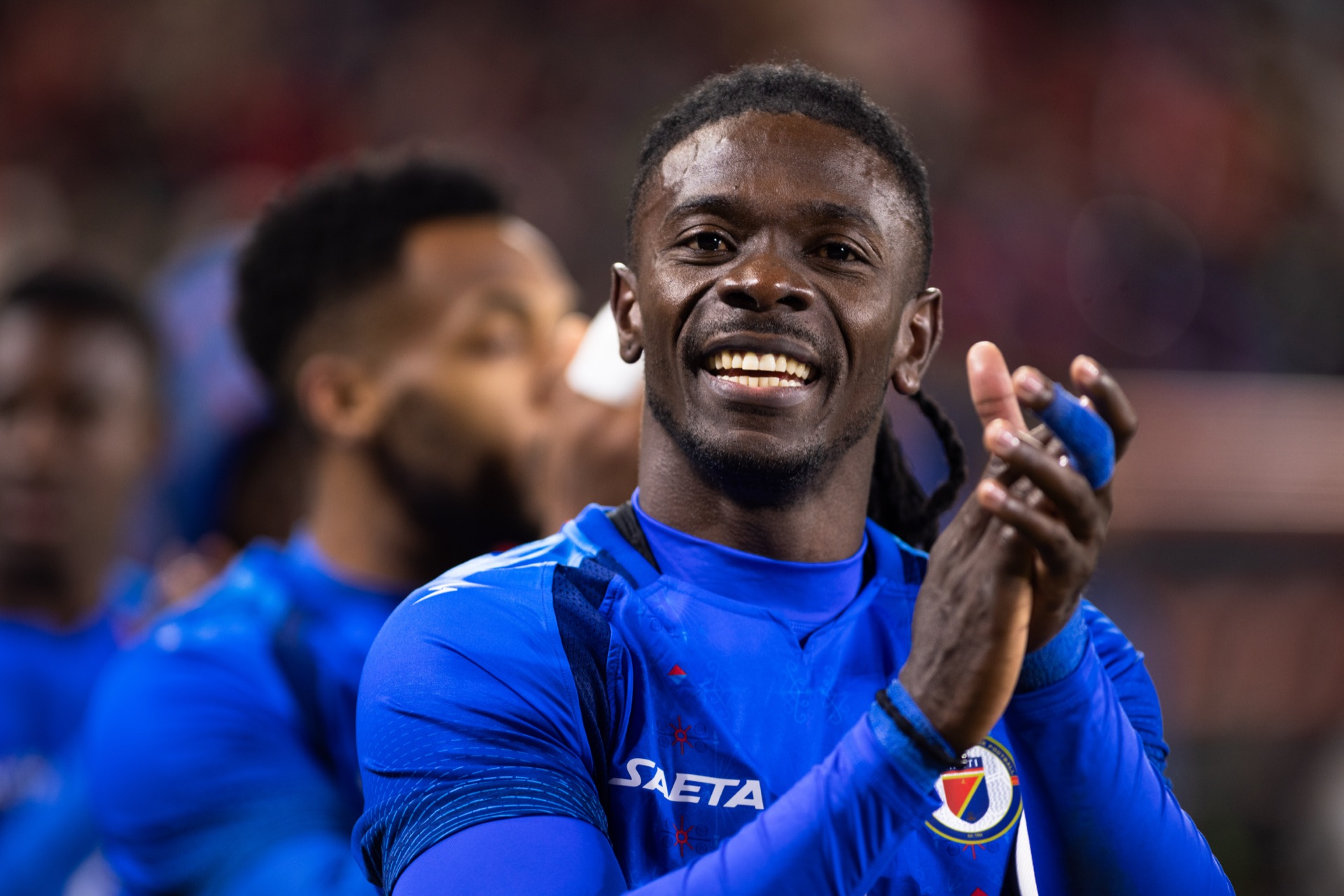 A Haiti player applauding the supporters after the whistle, grinning, teammates out of focus beside him