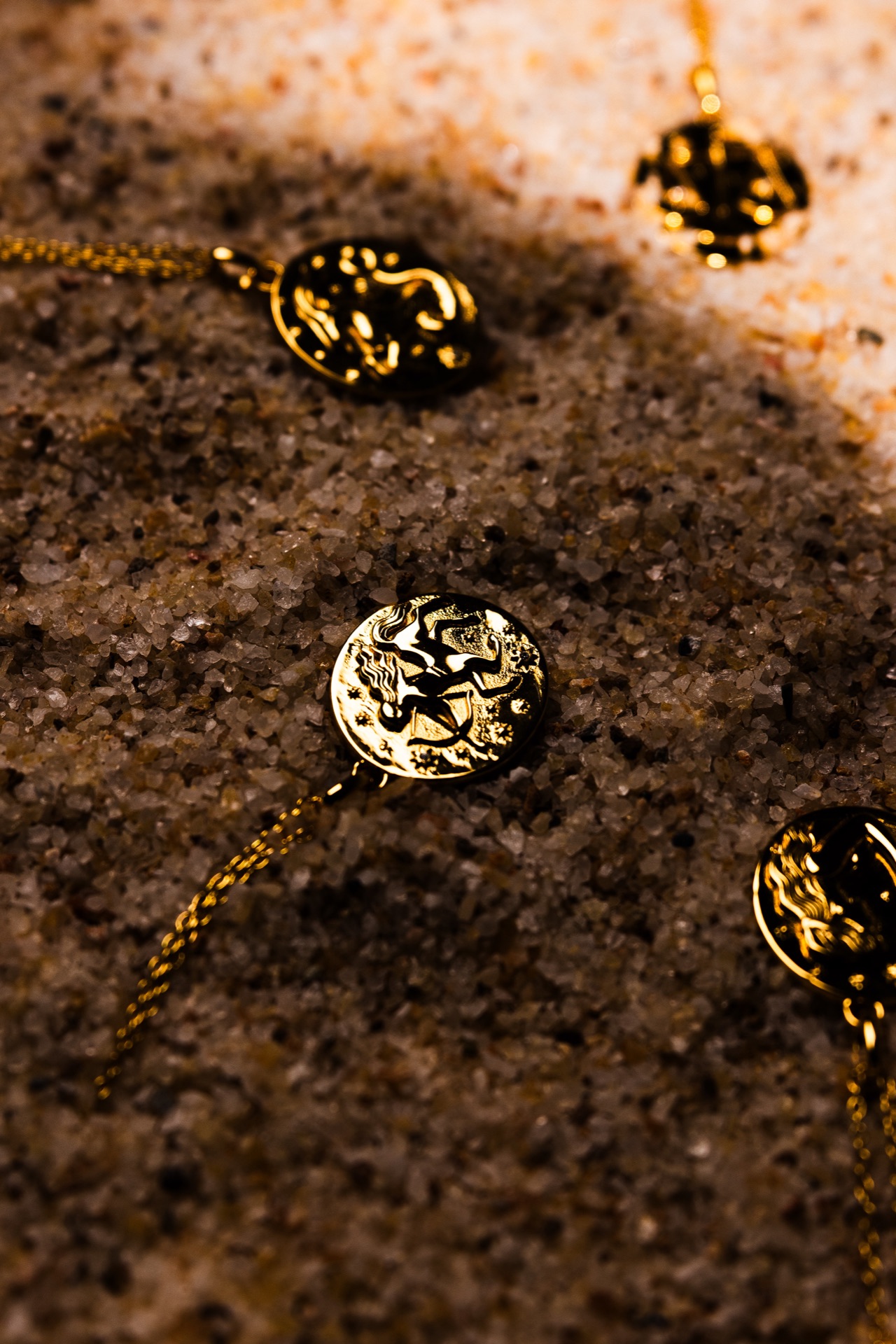 Gold and blackened zodiac pendants half-buried in dark wet sand, lit hard from one side