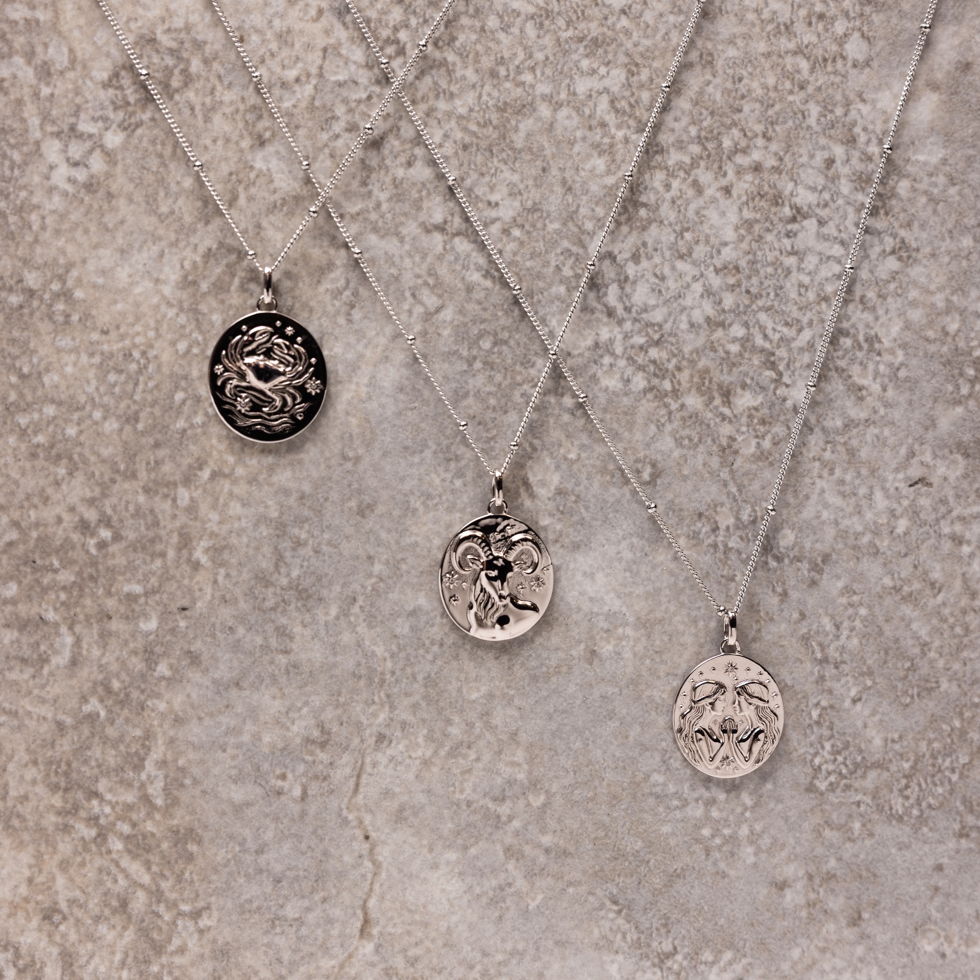 Three silver zodiac pendants laid out on a grey concrete slab, chains trailing across the stone