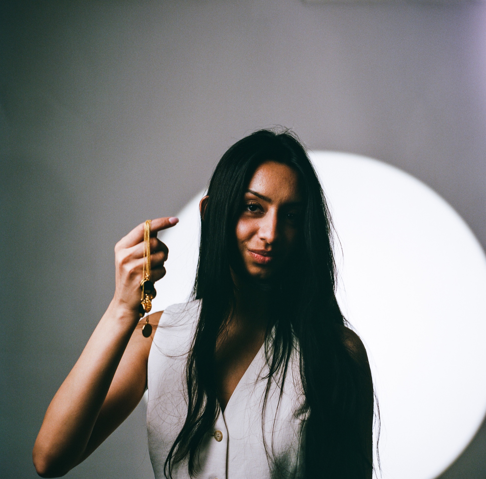 Model in a white shirt against a grey backdrop with a circle of light behind her, holding a gold pendant chain