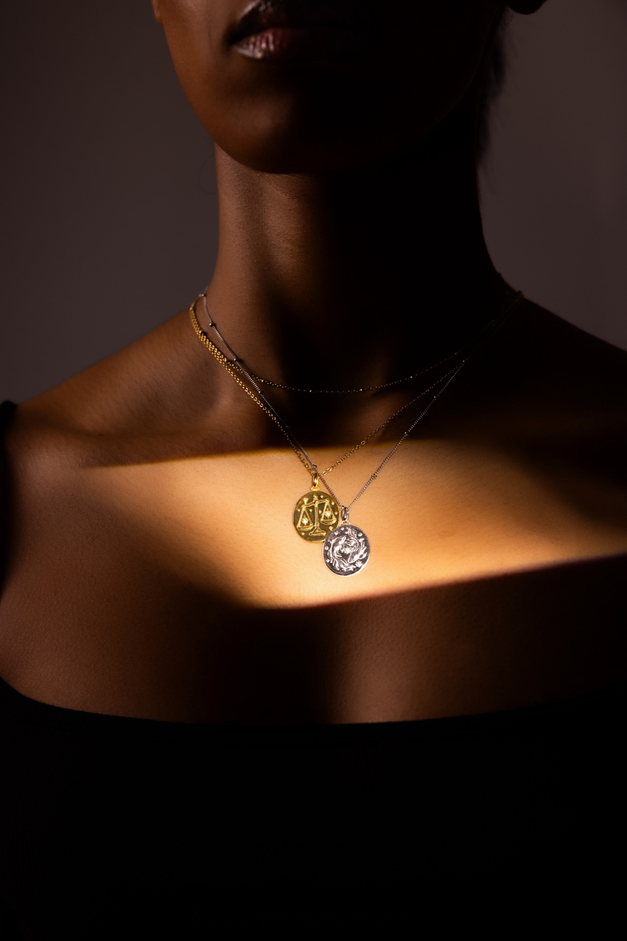 Low-key frame of a model's shoulder and collarbone, two gold pendants catching the only light in the shot