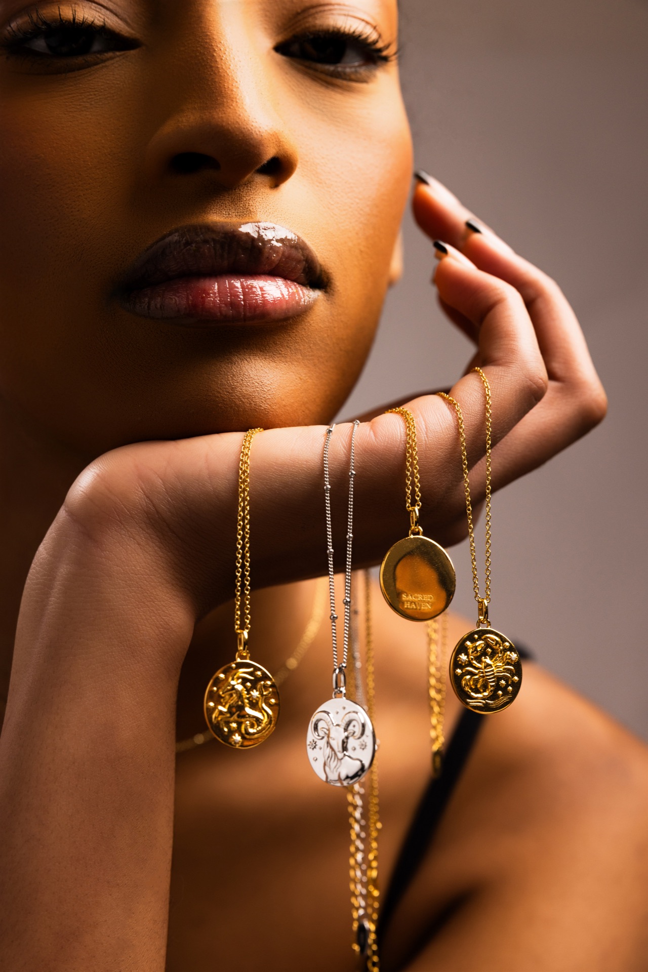 Model's face half out of frame, hand raised to her jaw, wearing stacked gold and silver zodiac pendants and bracelets