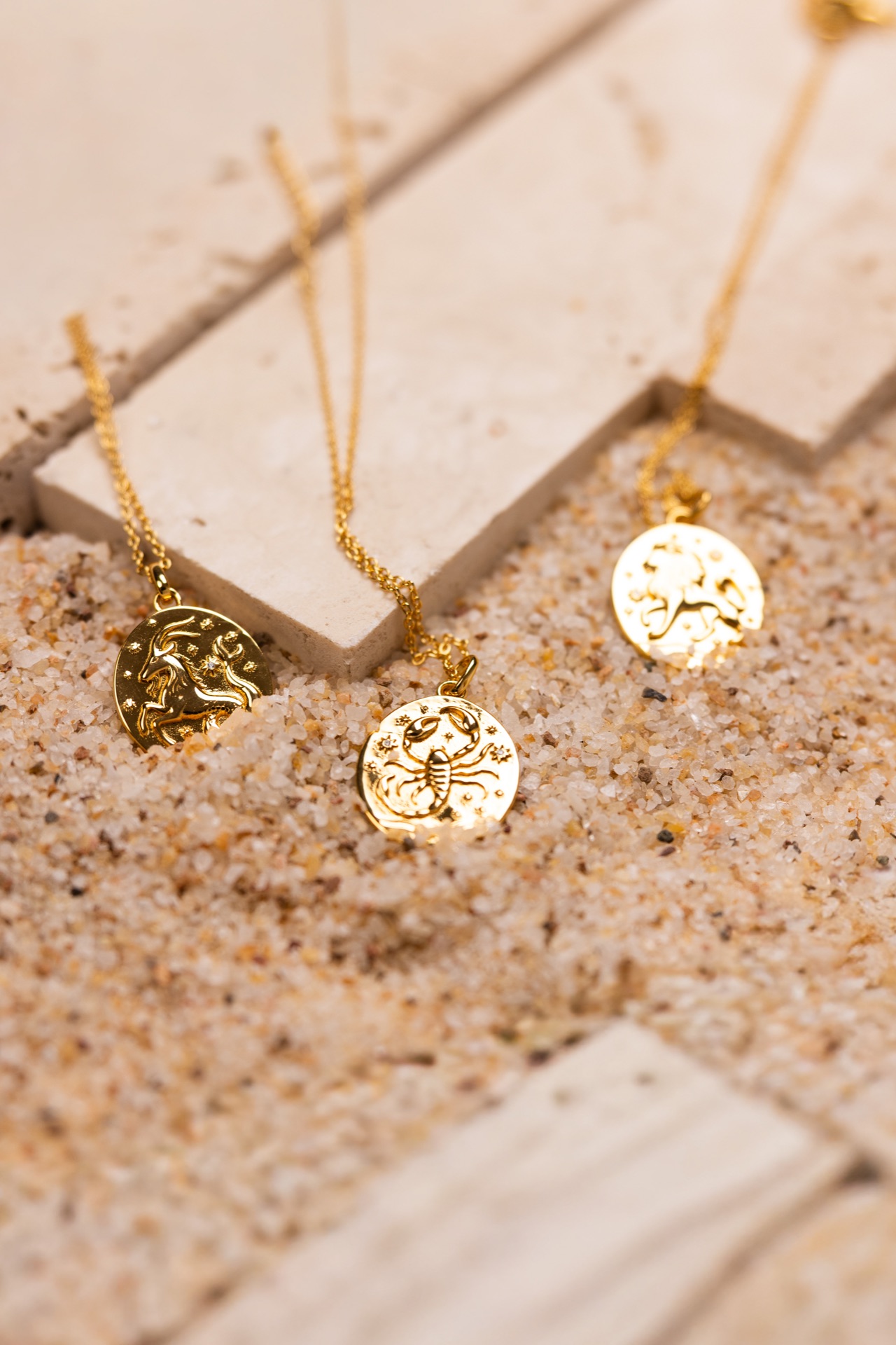 Gold zodiac pendants draped over travertine blocks set into sand, in warm directional light