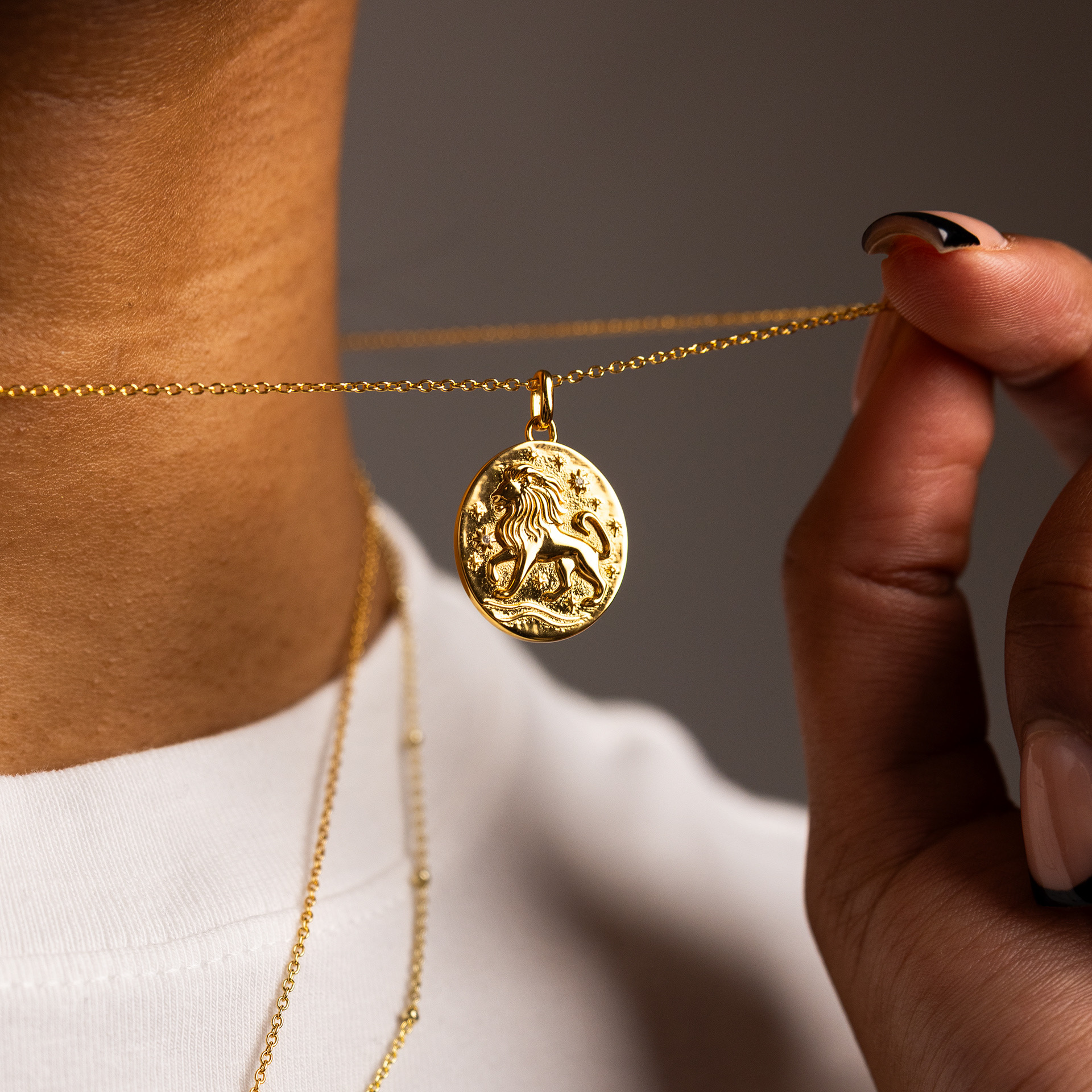 A hand lifts a gold lion pendant away from the chain at a model's neck, worn over a white tee