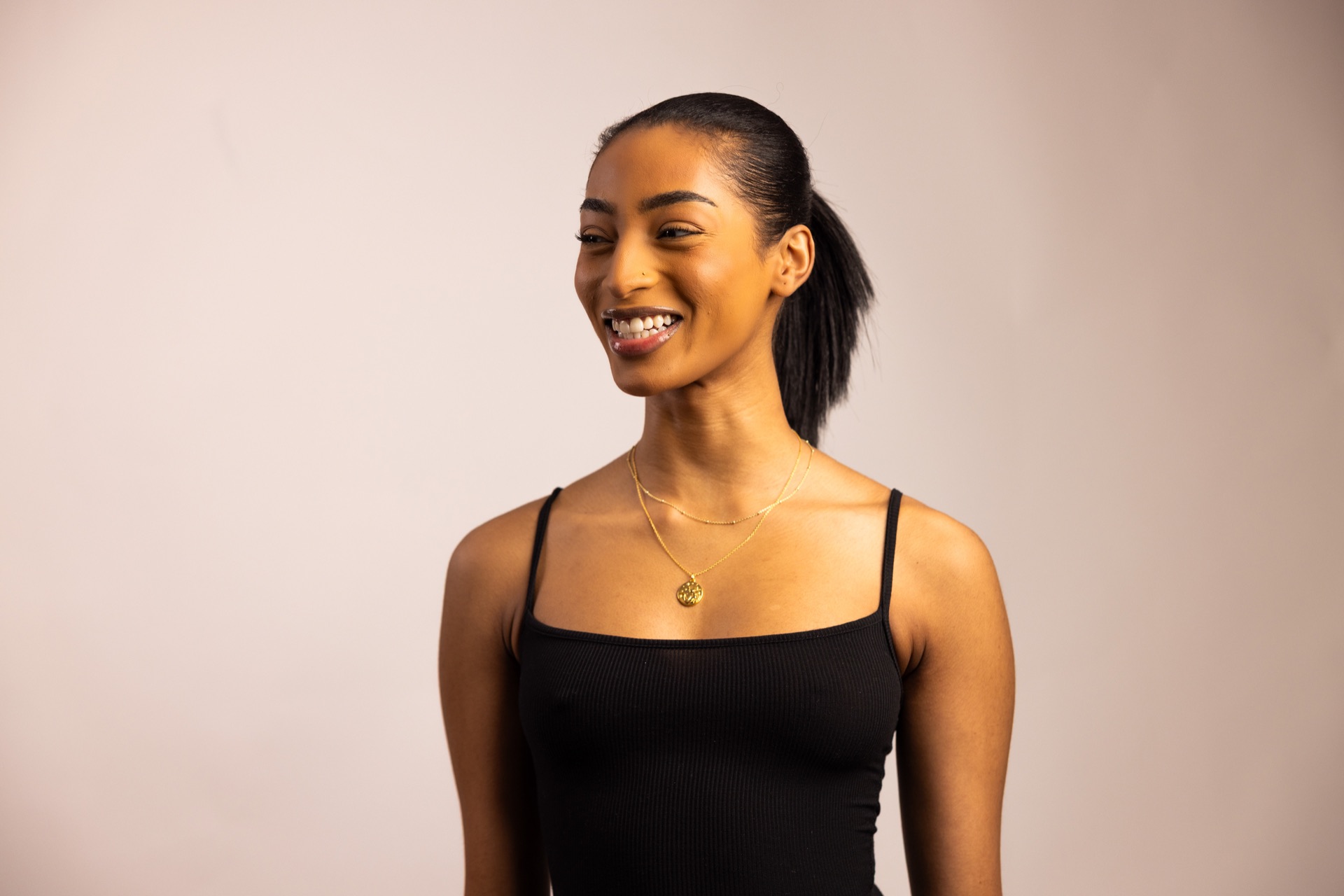 Model laughing in a black tank top against a warm studio wall, a single gold pendant at her neck