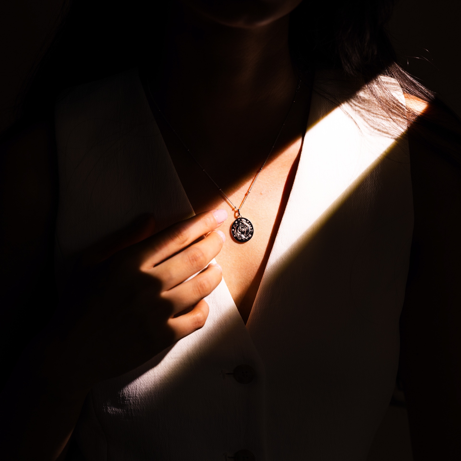 A shaft of daylight falls across a model's chest and hand as she holds a silver zodiac pendant against a white waistcoat