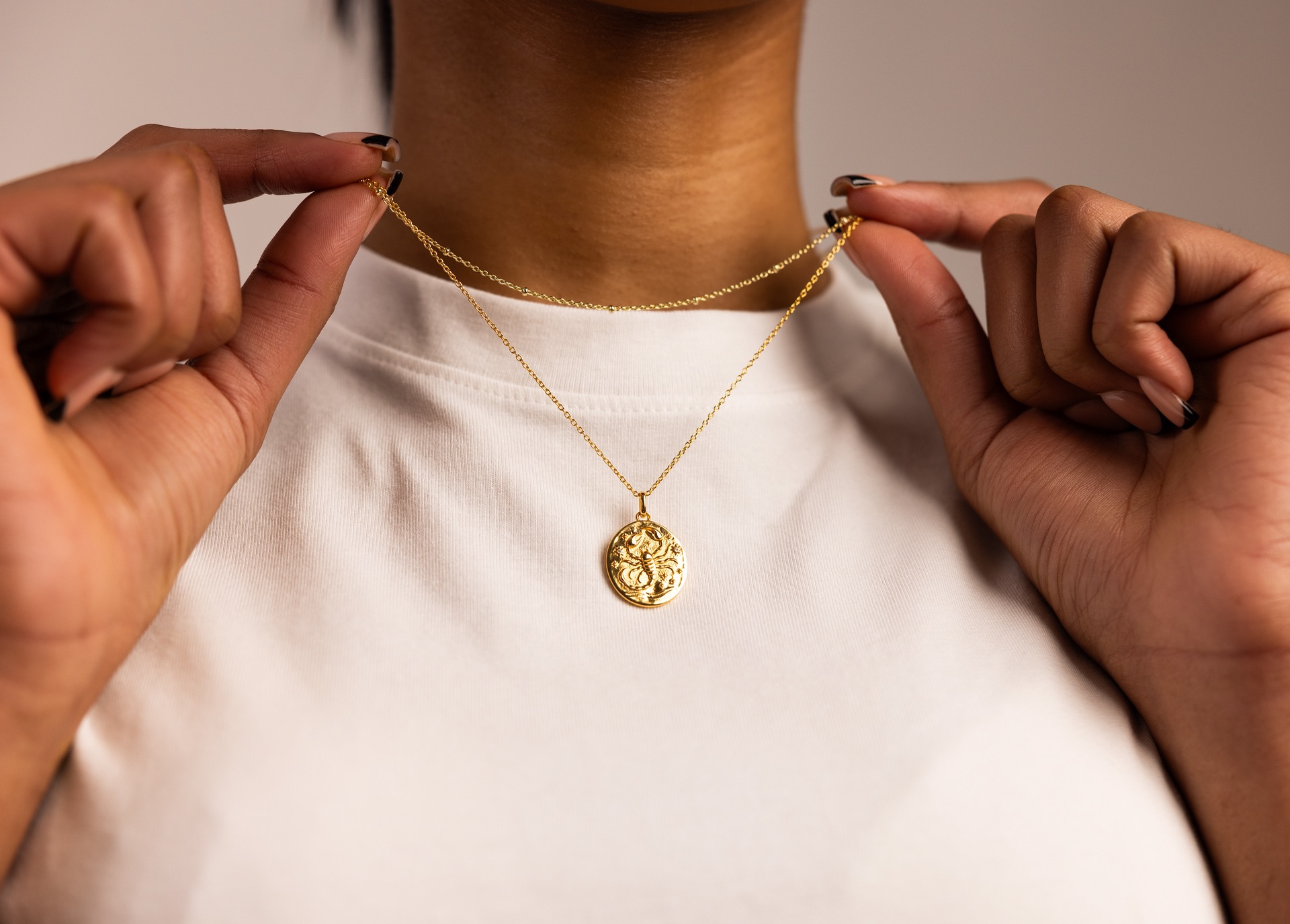 Two hands lift a fine gold chain at the collar of a cream top, a zodiac pendant resting below