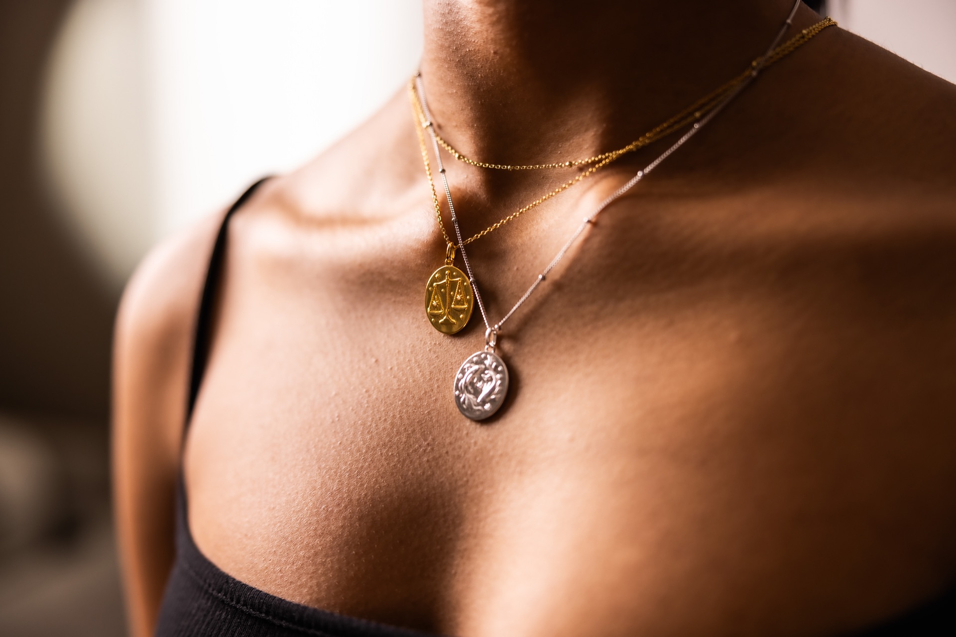 Hard warm light across a model’s collarbone and shoulder, a gold and a silver zodiac pendant hanging together