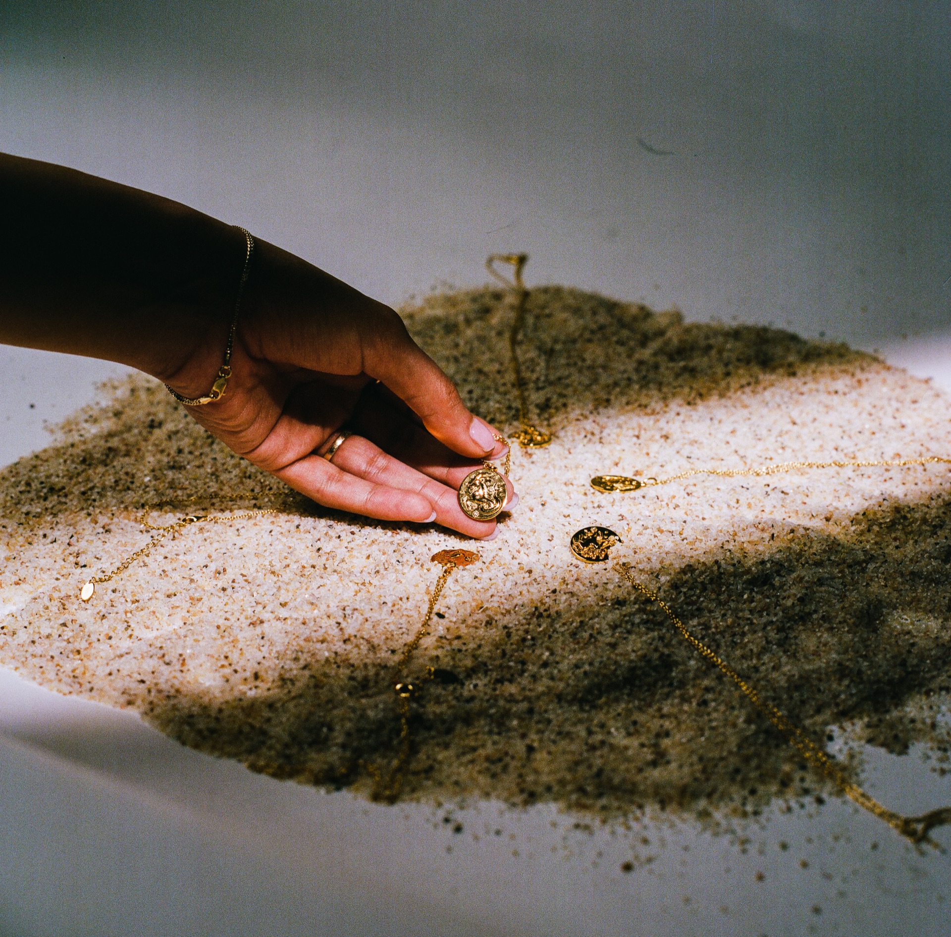A hand sets a gold pendant into wet sand, chains scattered around it