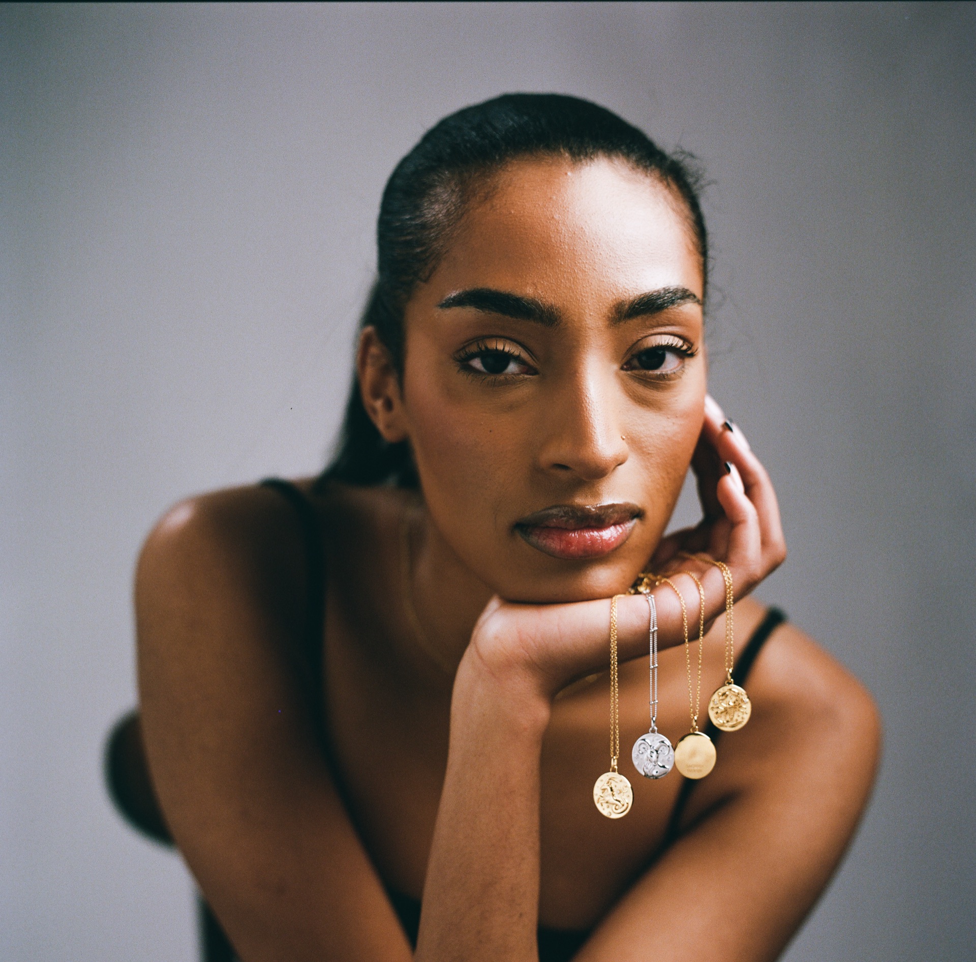 Studio portrait of a model resting her chin on her hand, three small zodiac pendants layered at the neck