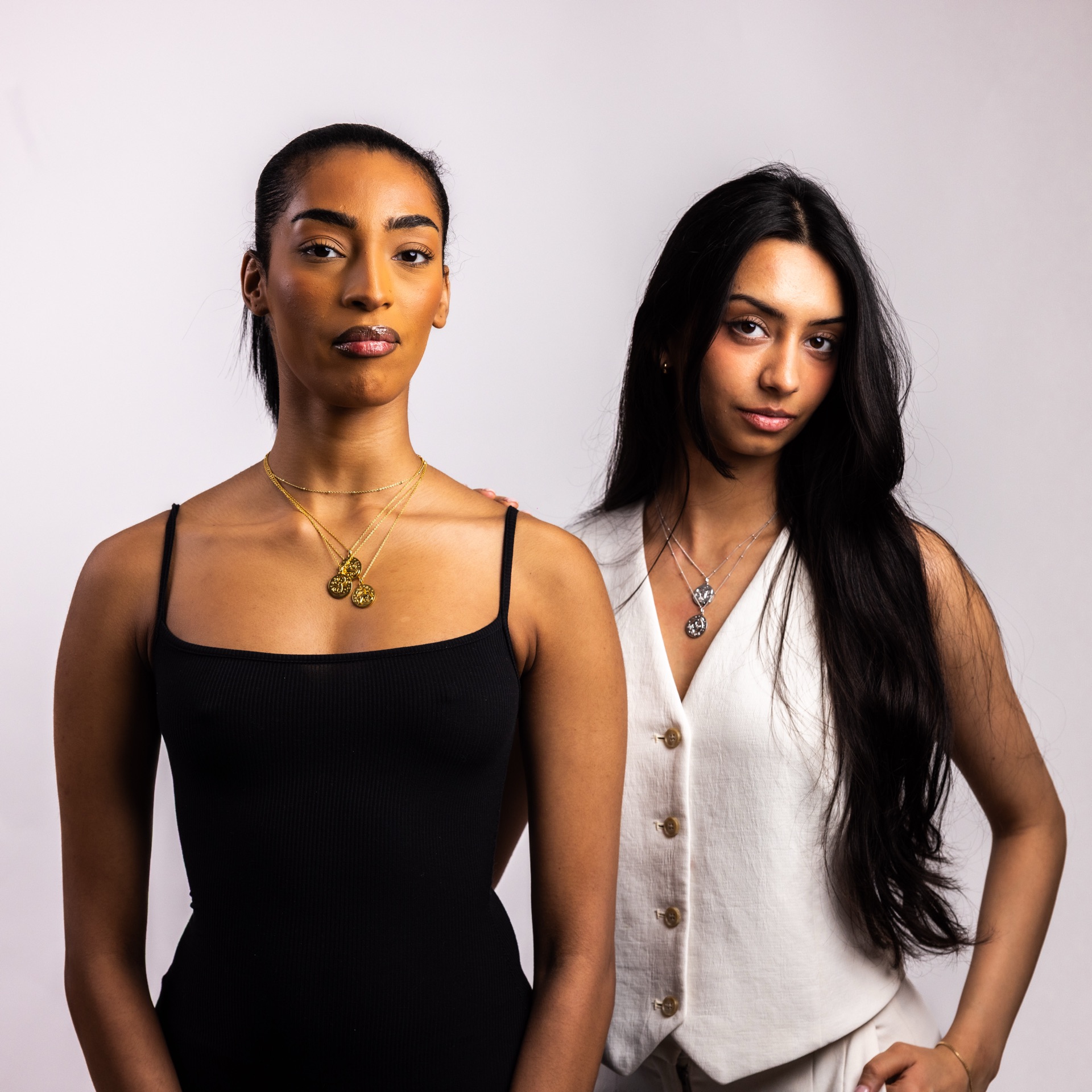 Two models, one in a black slip dress and one in a white waistcoat, wearing layered Sacred Haven zodiac pendants against a pale studio backdrop