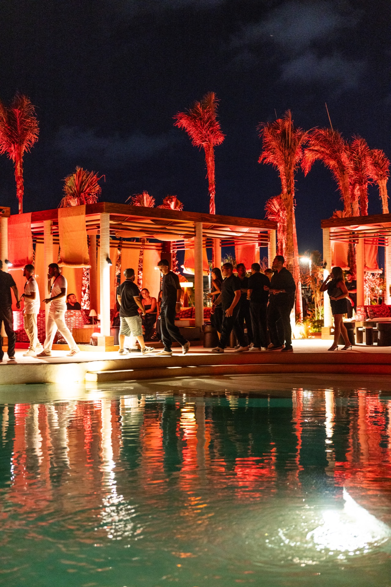 Guests along the poolside under lit palms before the sets begin