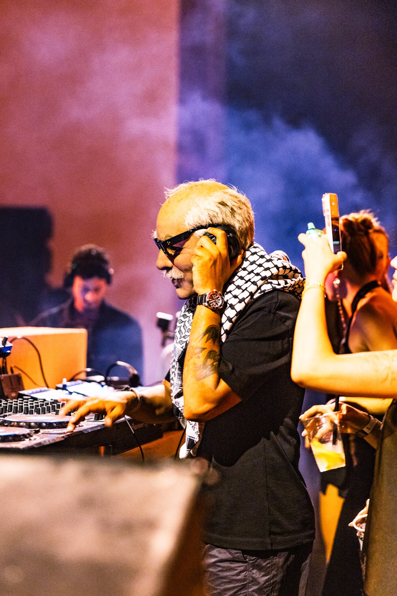 A hand to the headphones at the booth, a second DJ behind in the haze