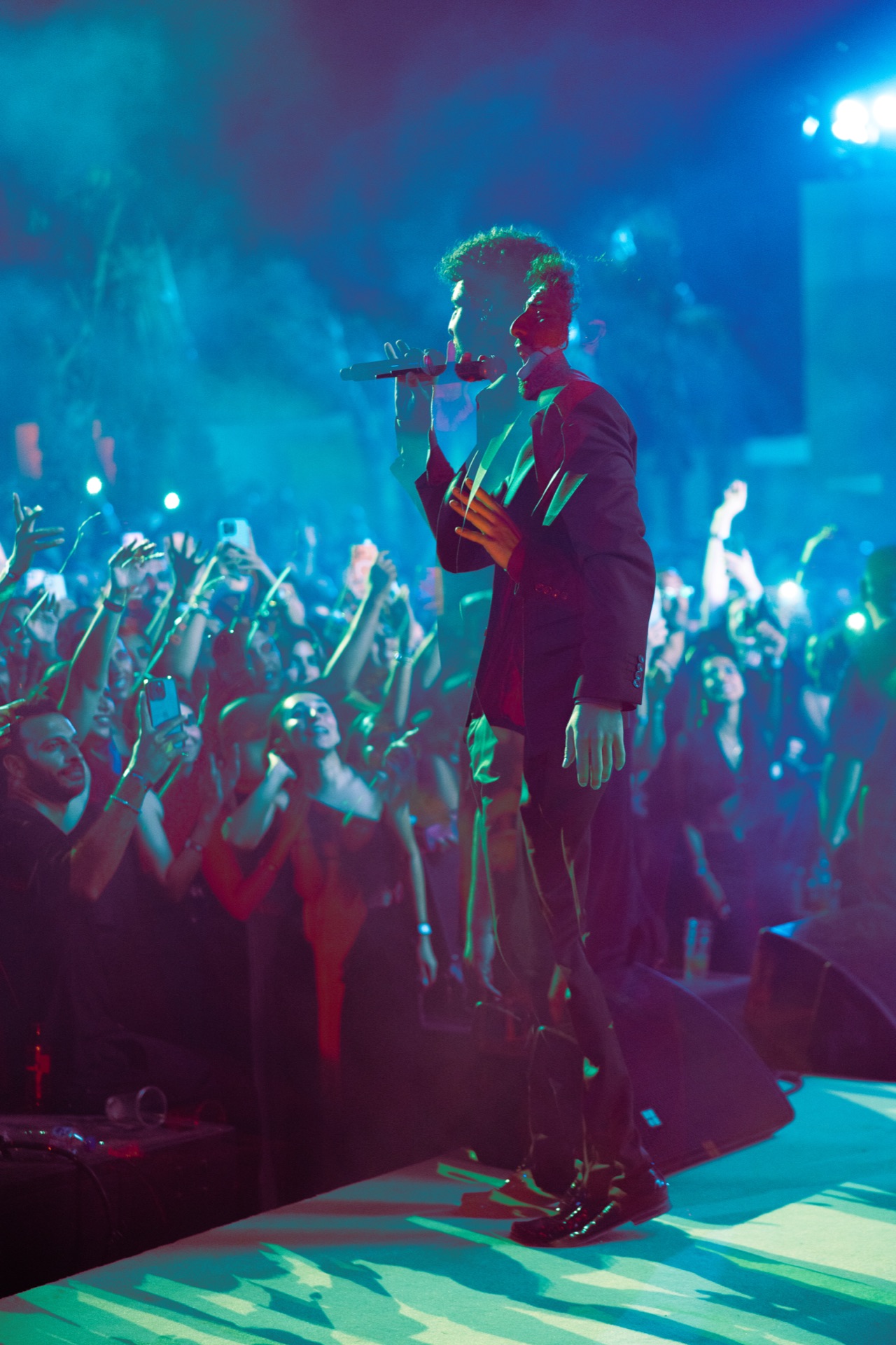 Full-length on the stage lip, the crowd stretching back under green light