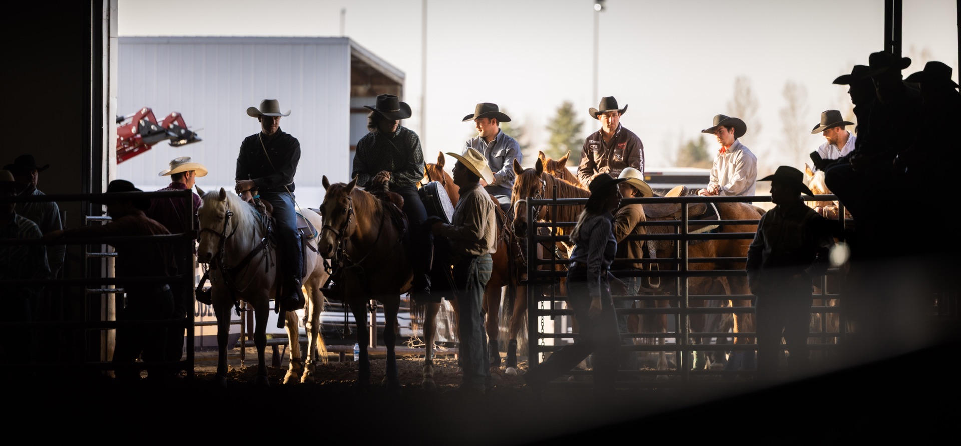 Riders wait on horseback in a lit doorway at the back of the grounds, seen through the pens