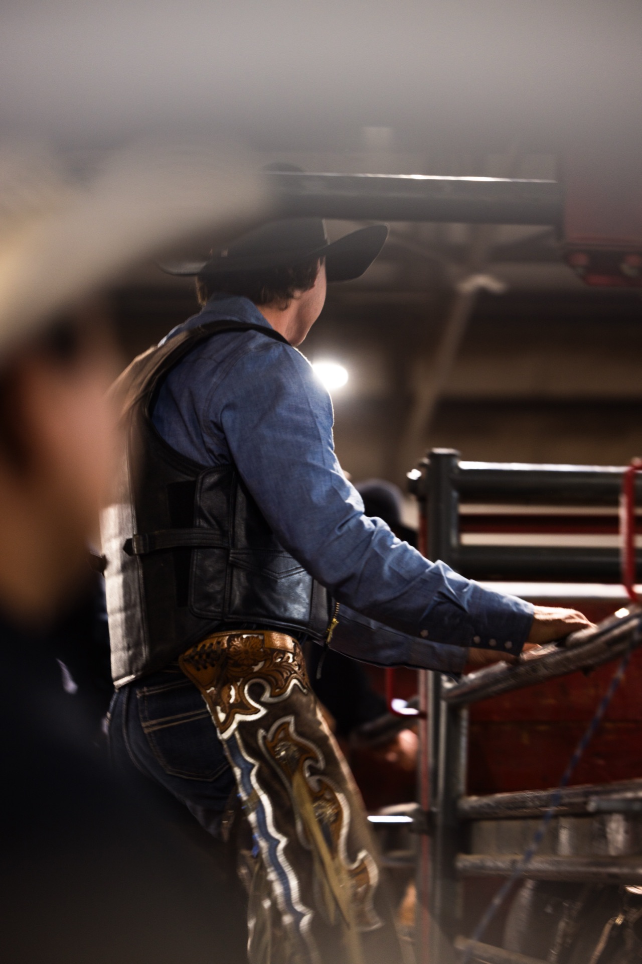 A rider in a protective vest and tooled chaps leans on the chute rail, back to camera