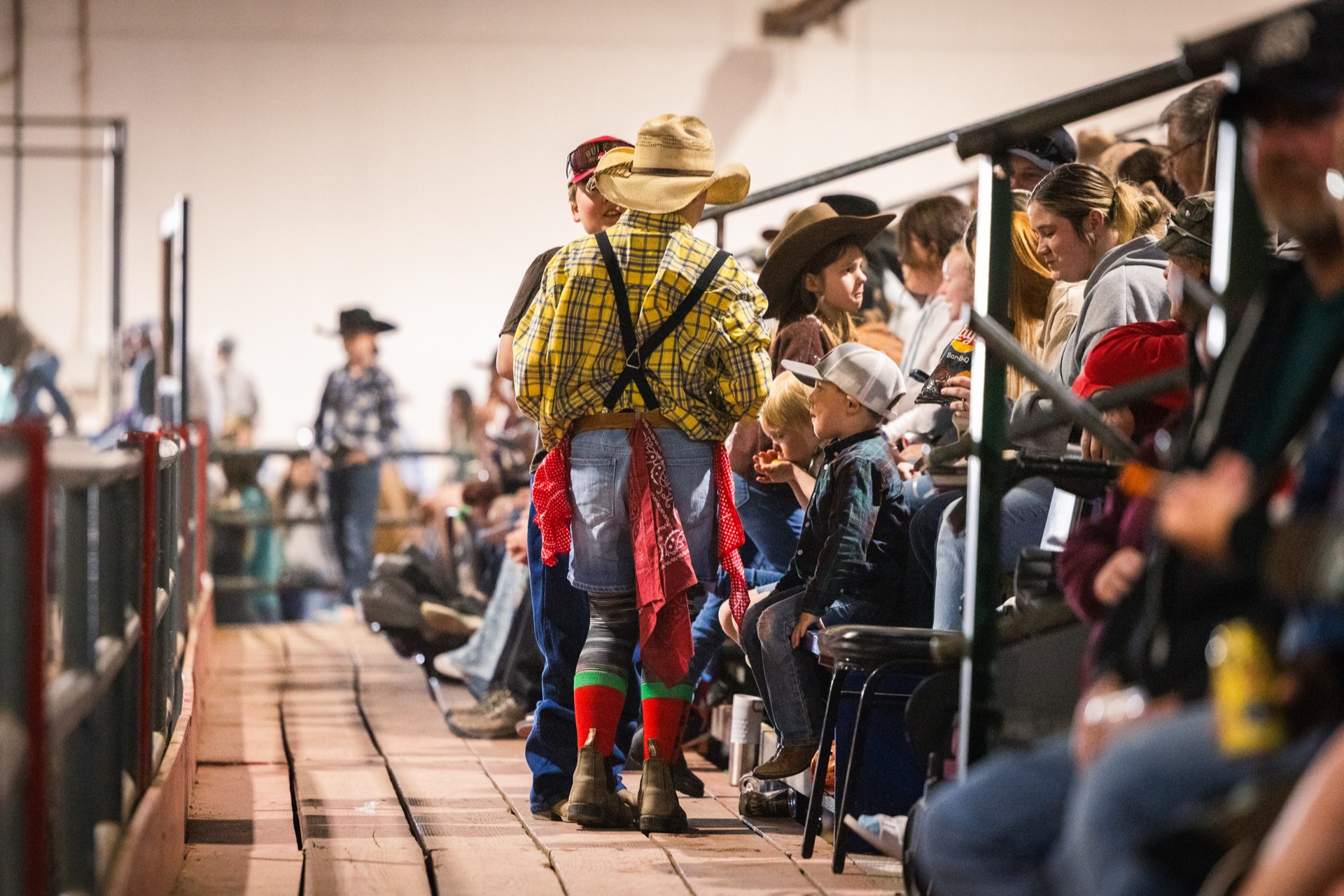 A rodeo clown in a straw hat and suspenders walks the boards past families in the stands