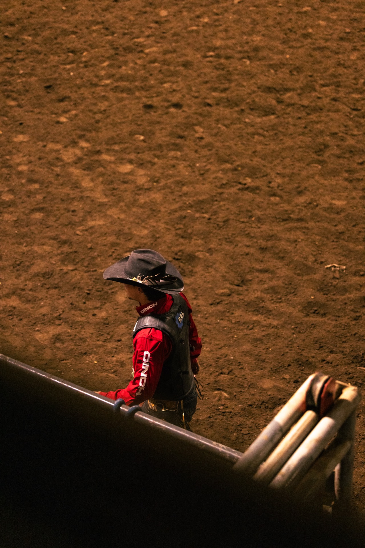 A competitor in a red shirt and protective vest walks out across the dirt, seen over the arena rail