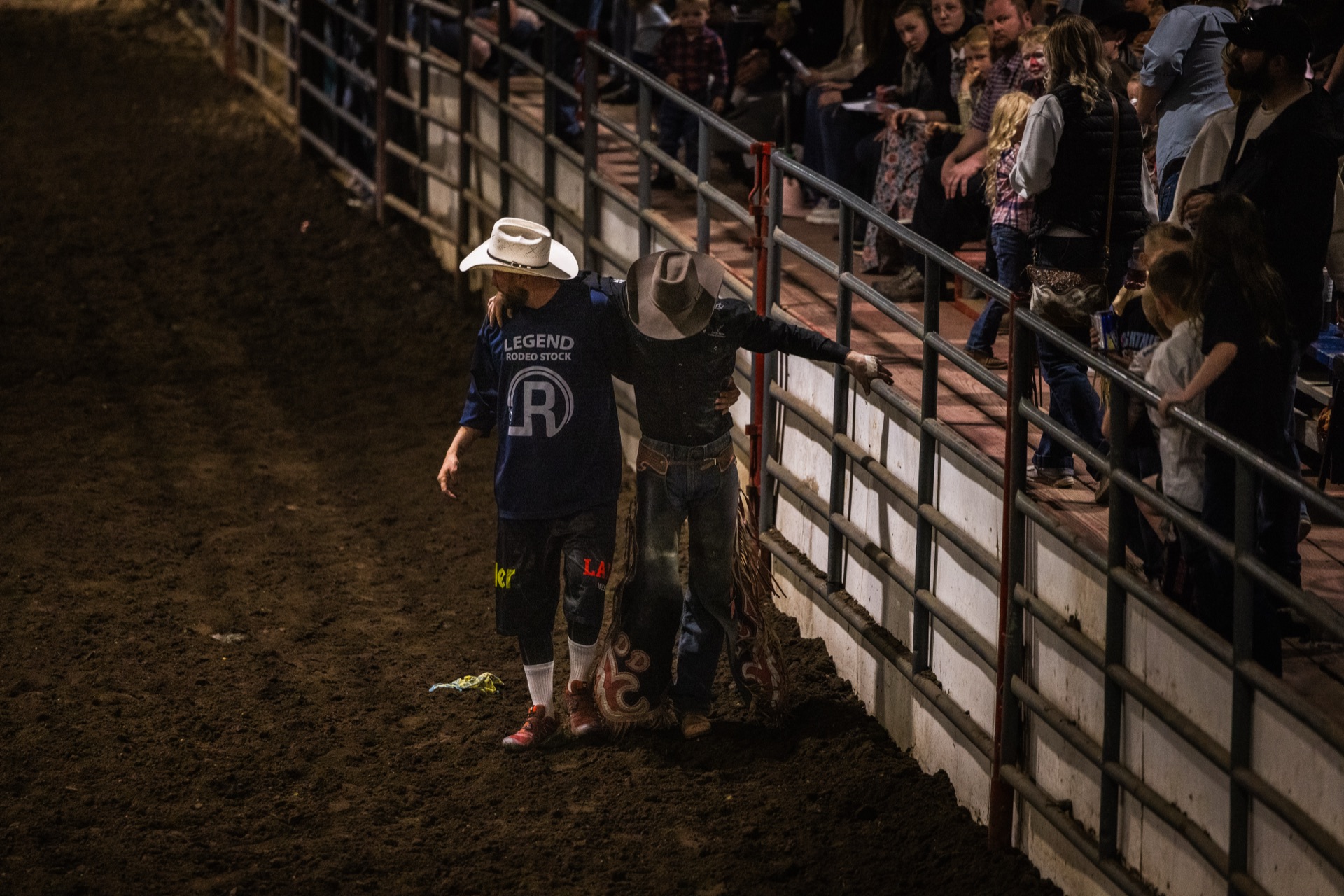 A helper walks an injured or spent rider along the arena rail as the crowd watches from the stands