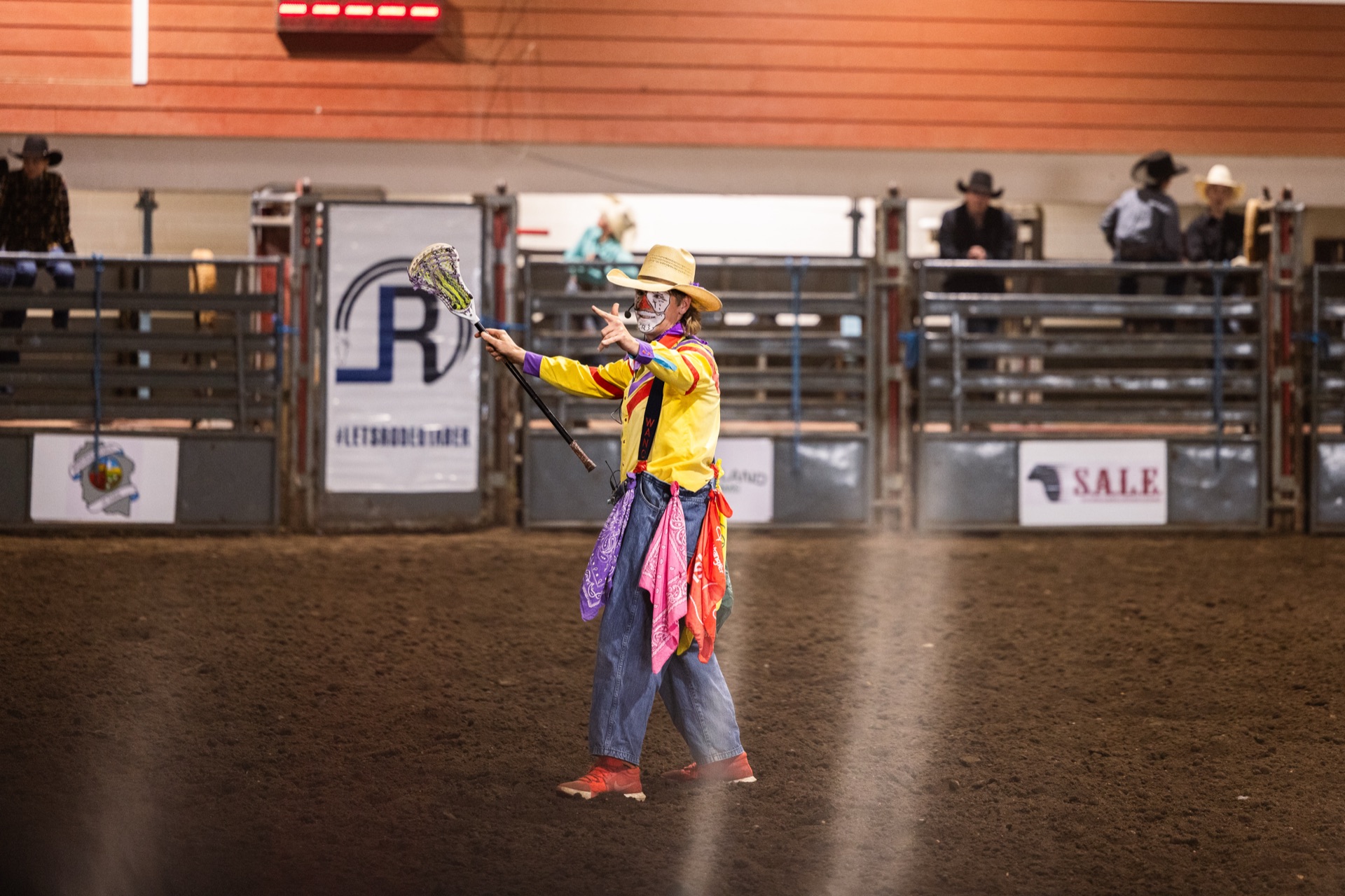 A rodeo clown in a yellow shirt and patchwork coat gestures to the crowd from the middle of the arena