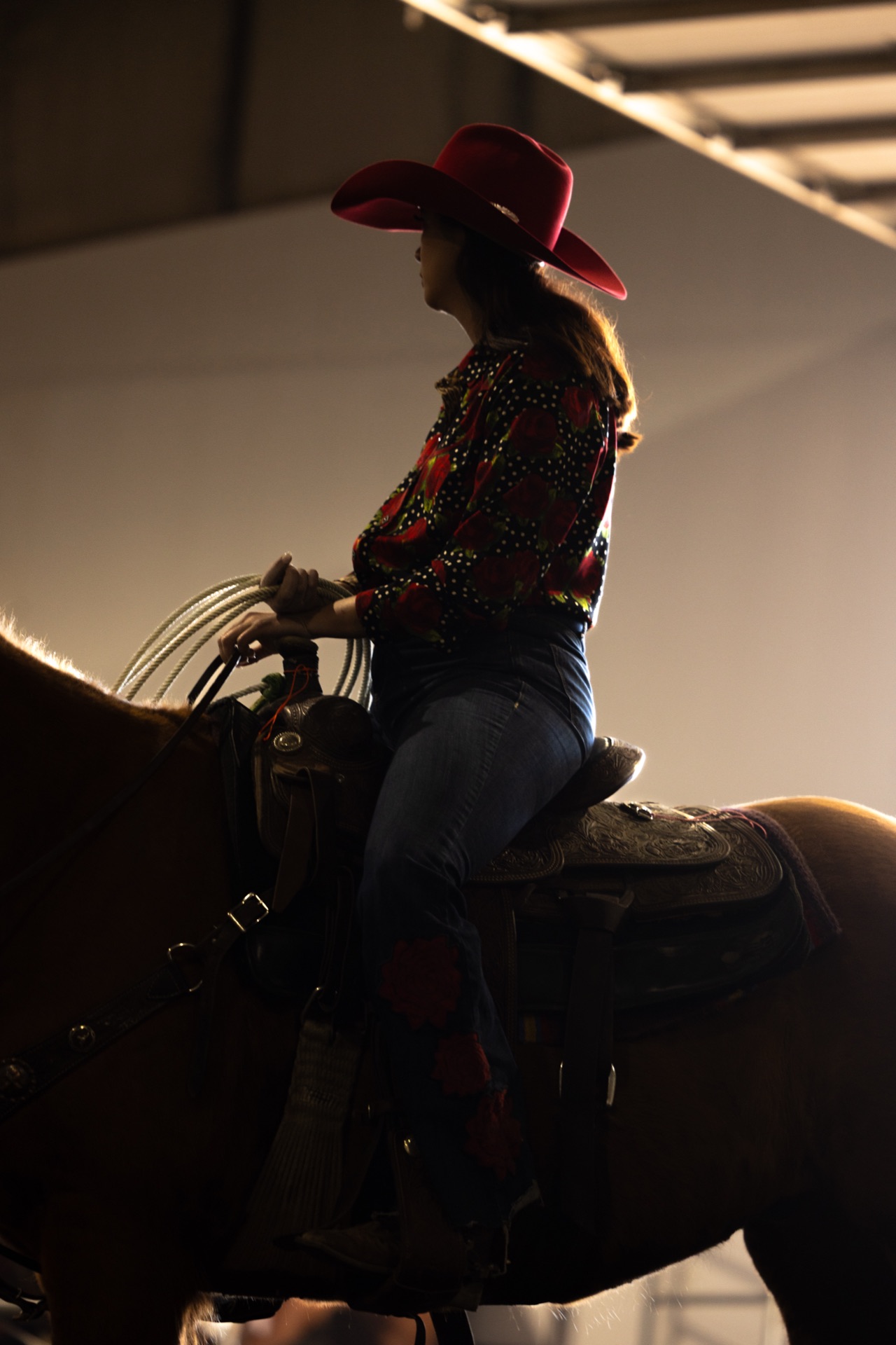 A woman in a red hat and floral shirt sits her horse in the warm-up light, rope coiled at the saddle