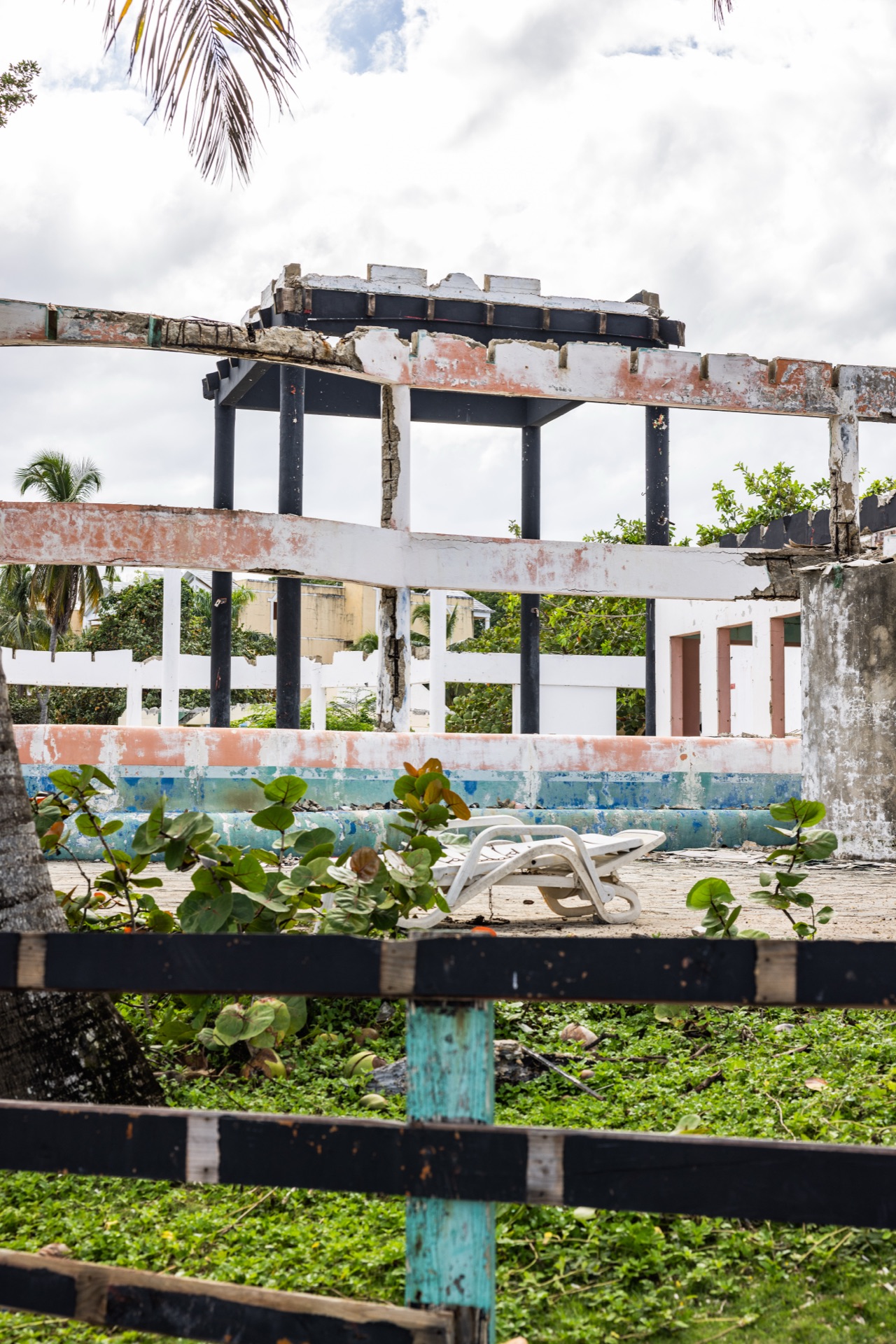 A gutted concrete building seen over a low fence, its roof stripped to the beams and a single plastic lounger left beside the drained pool