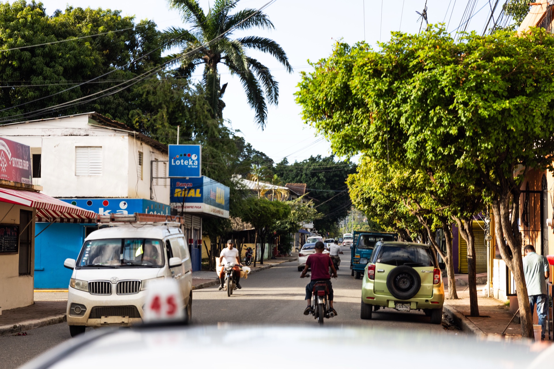 A main street through town — motorbikes, a parked minibus and a lottery sign under heavy trees