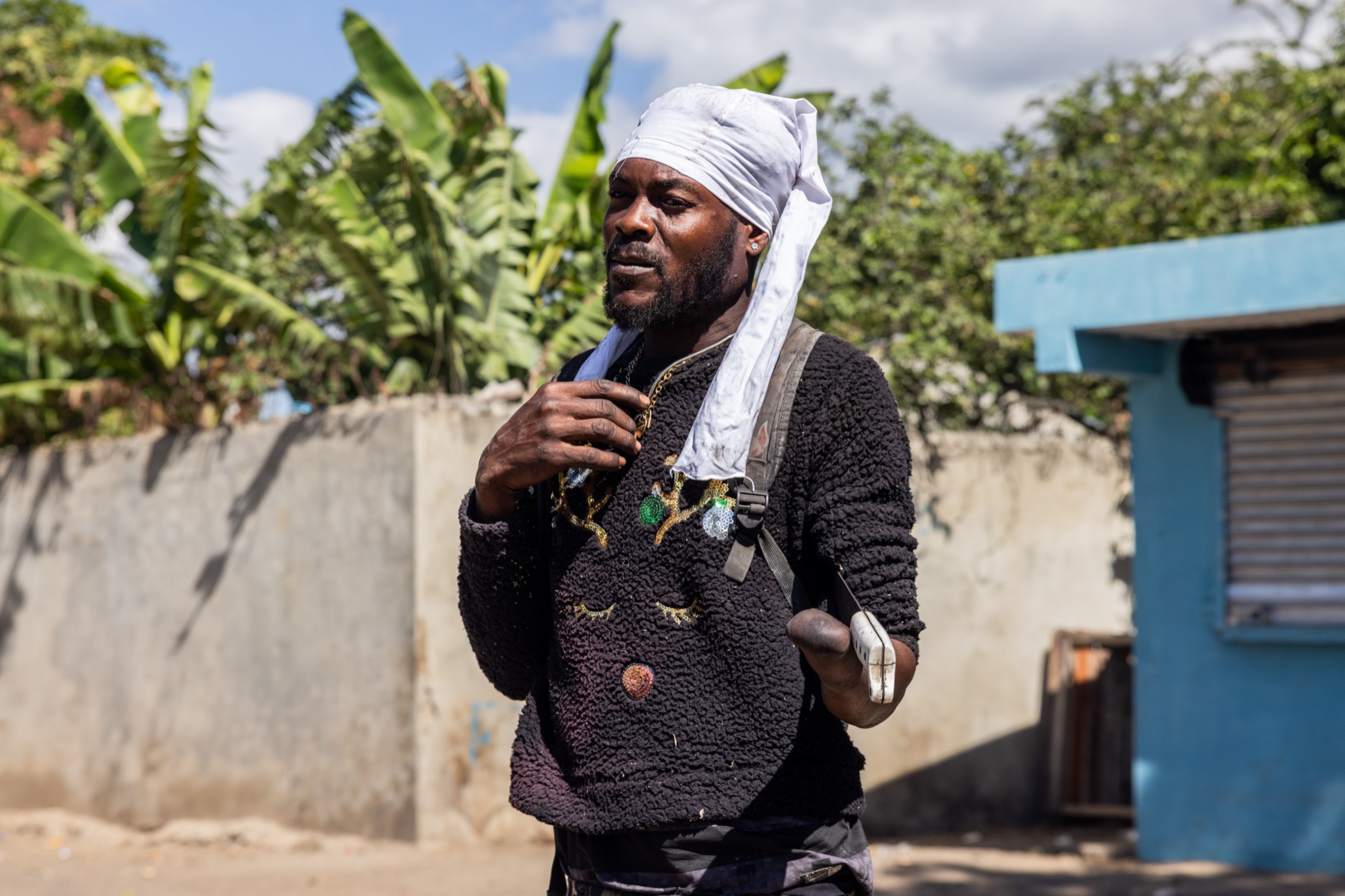 A man with a cloth wrapped around his head walks past a banana grove and a blue-painted building in hard midday sun