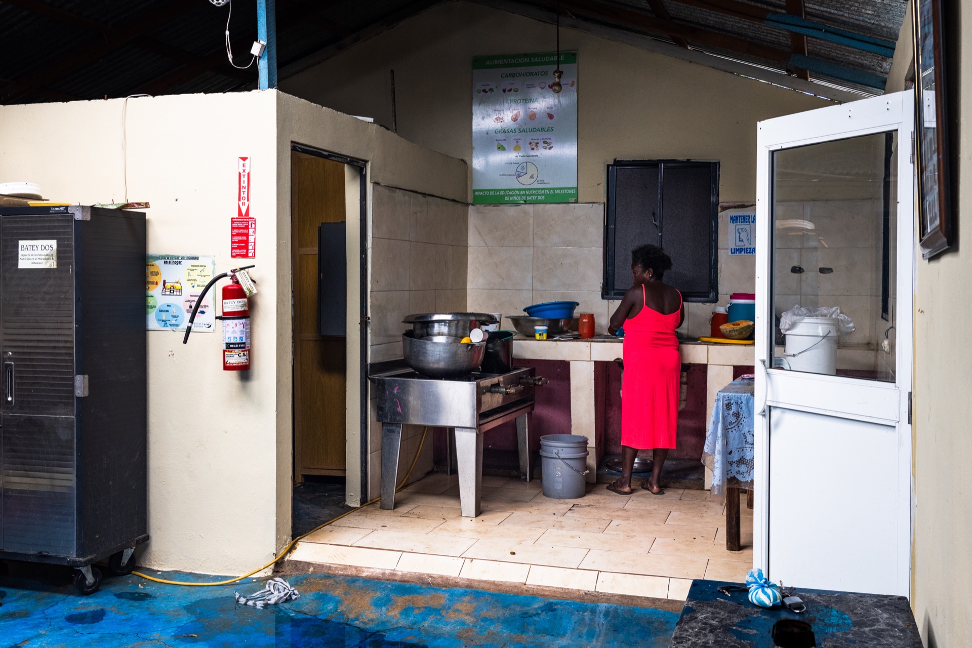 A woman in a red dress stands at the counter of a small tiled kitchen, a pot on the stove behind her