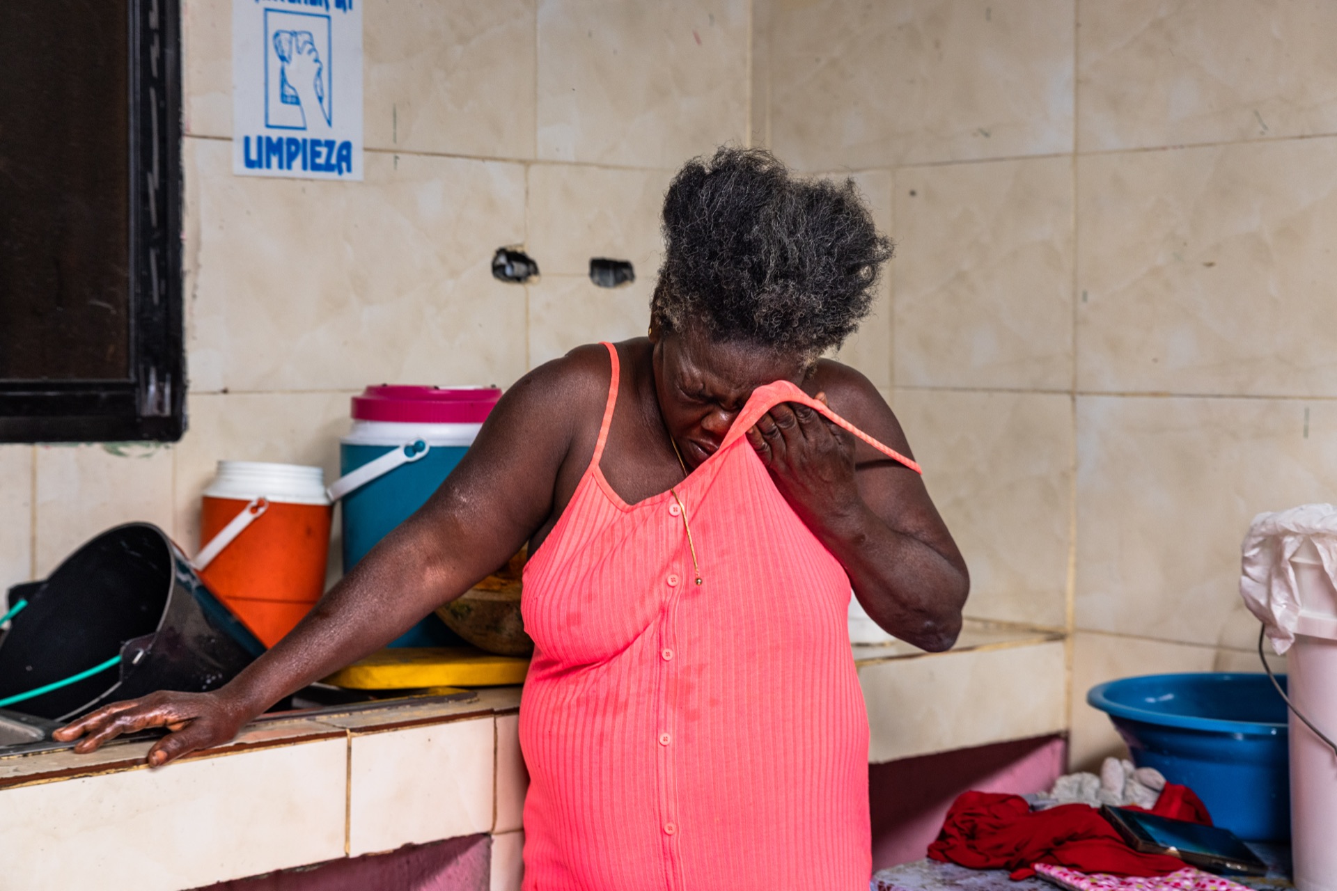 A woman in a coral dress leans on a tiled counter with one hand at her shoulder, beneath a printed cleaning notice