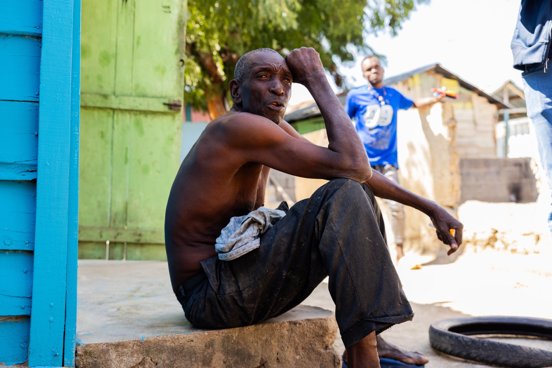 A shirtless man sits on a low step against a green and blue painted wall, resting his head on his hand as another man passes behind