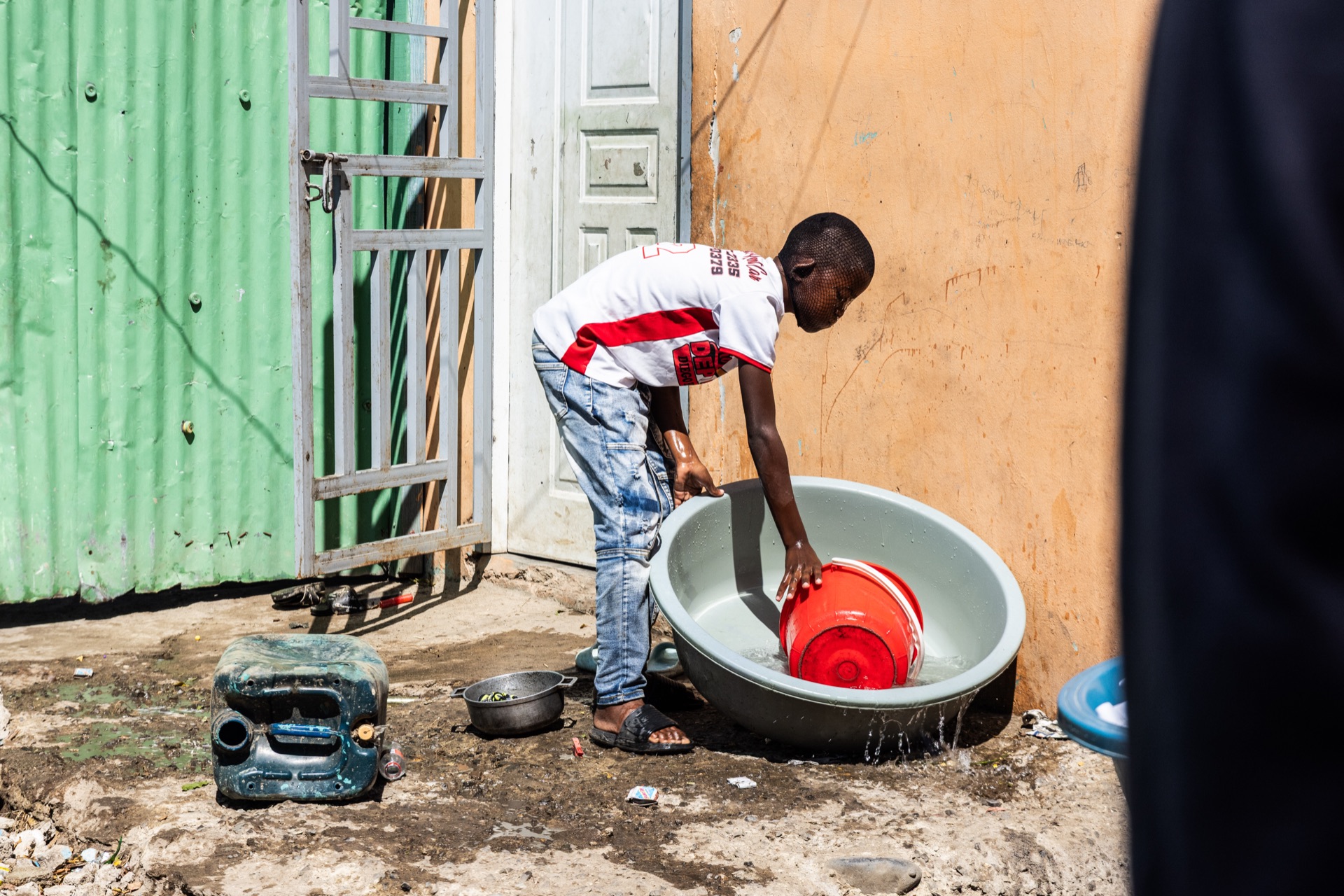 A boy bends over a large basin outside a corrugated-metal wall, washing up beside a jerrycan