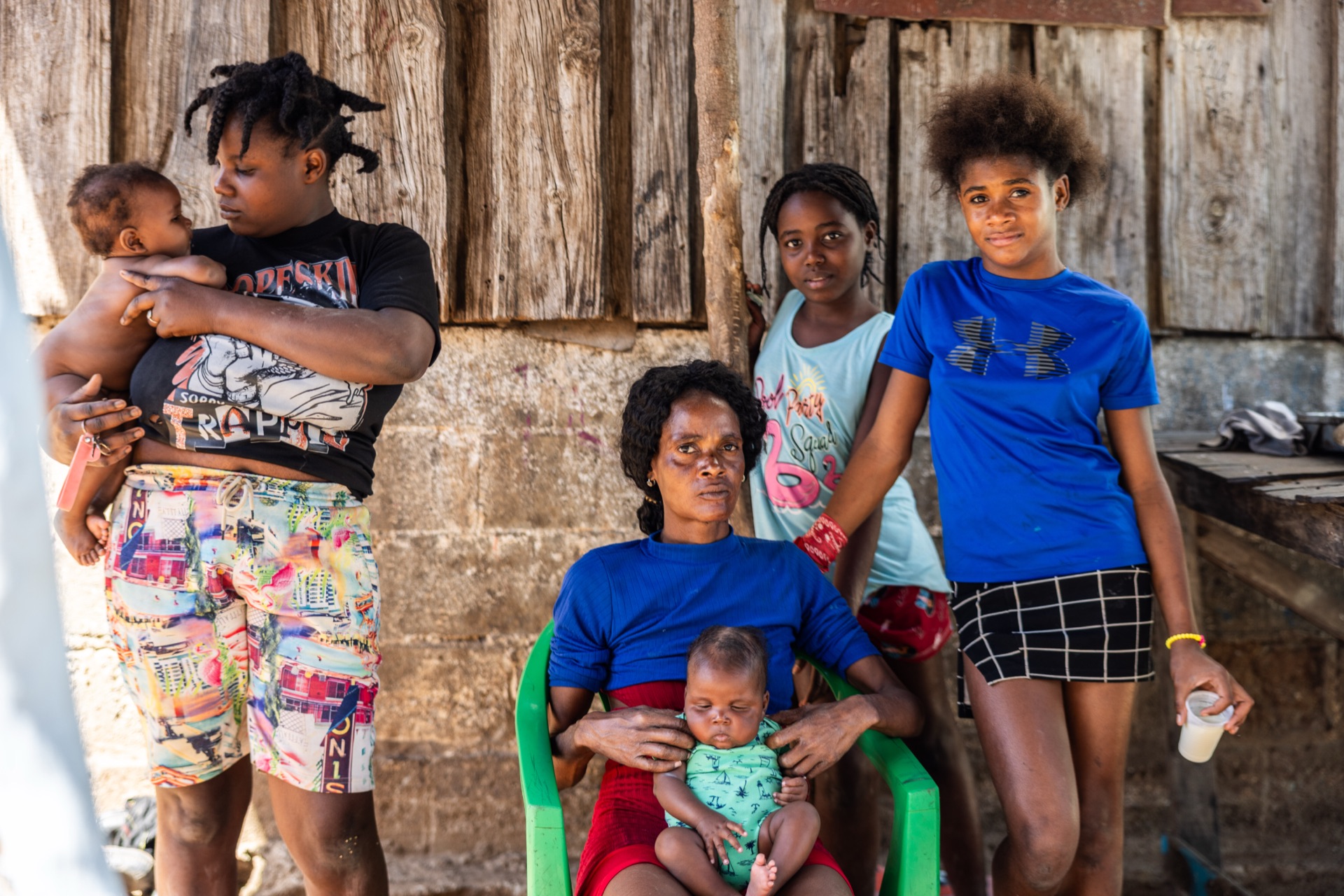 A family outside a weathered timber house — a young woman holding a baby, an older woman seated with an infant on her lap, and two girls standing alongside