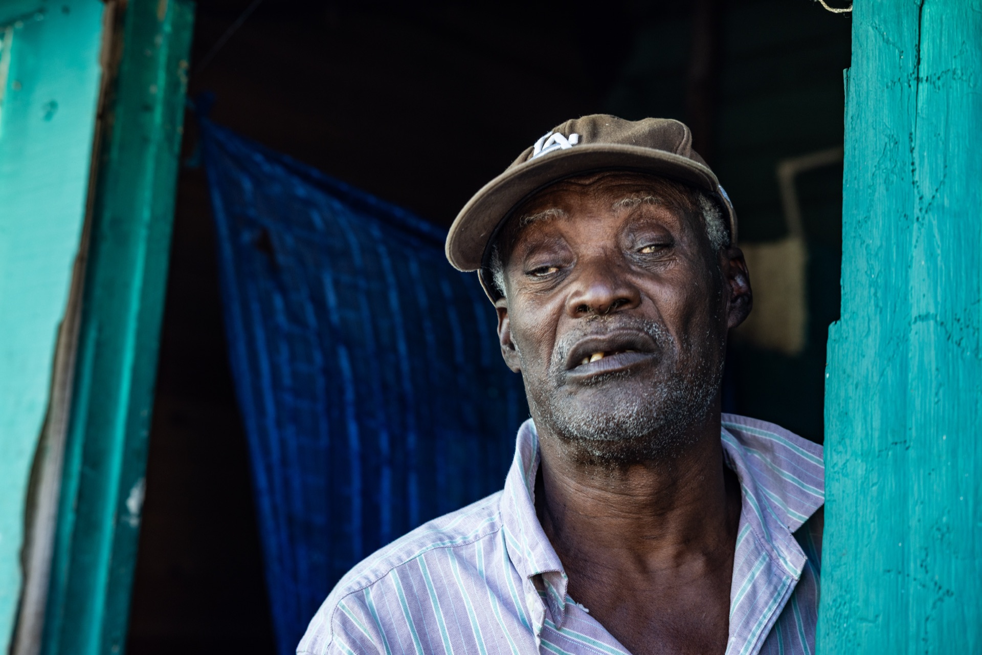 An older man in a cap and striped shirt stands in a turquoise doorway, mid-sentence