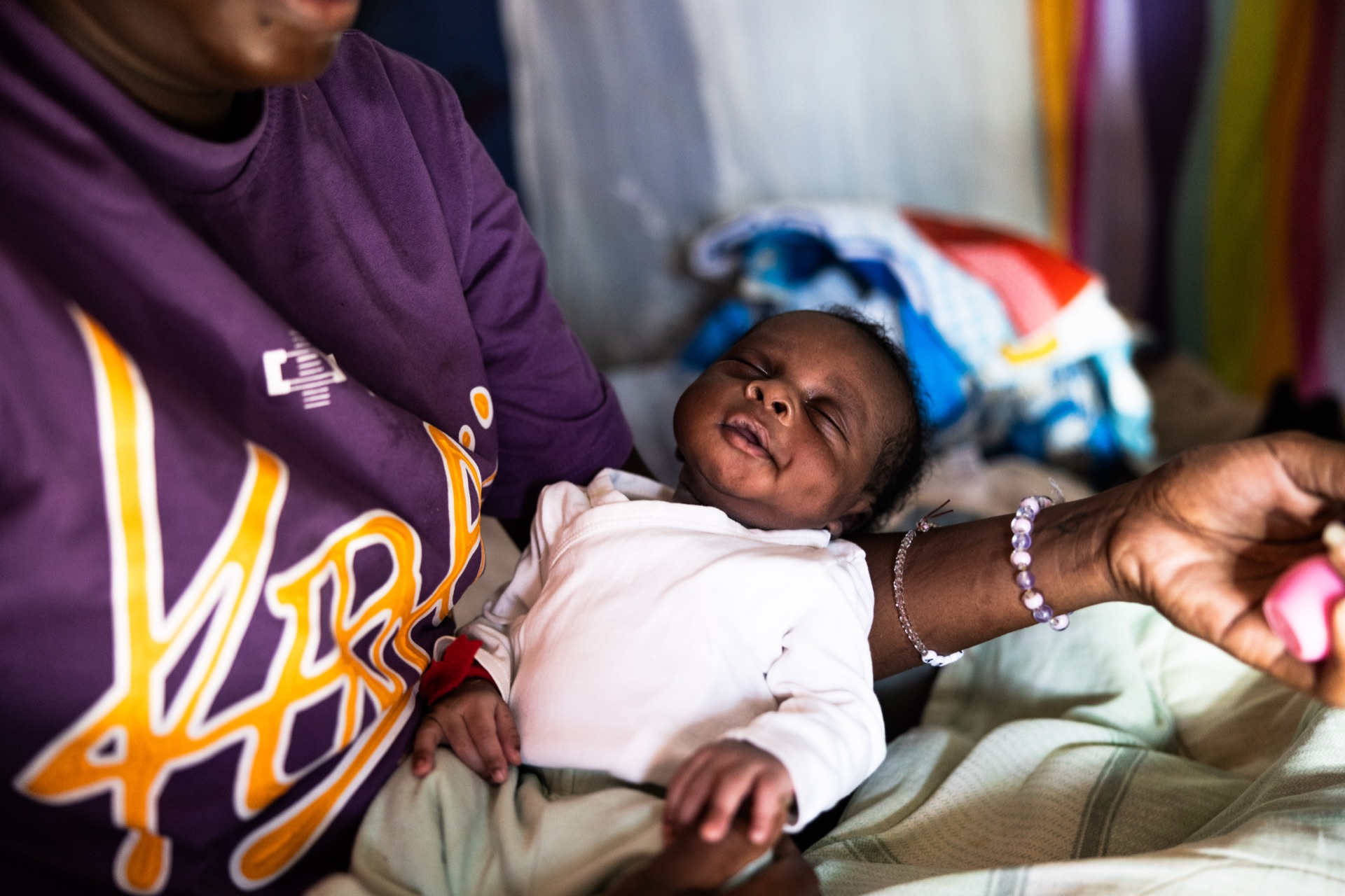 A newborn in a white top lies across an adult's lap indoors, eyes screwed shut, a beaded bracelet on the wrist reaching toward them
