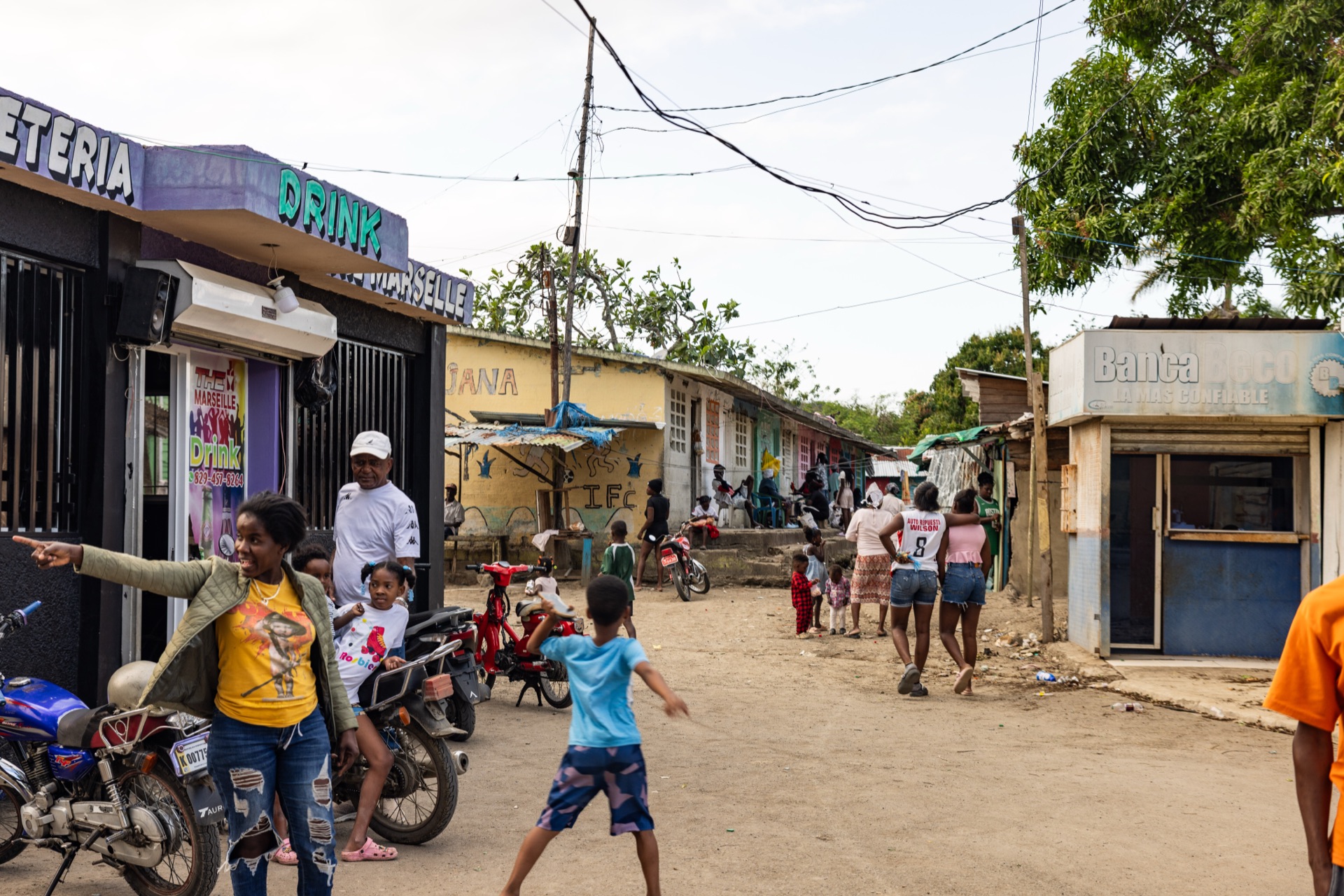 A busy street of small shopfronts, children playing in the road and motorbikes parked along the kerb